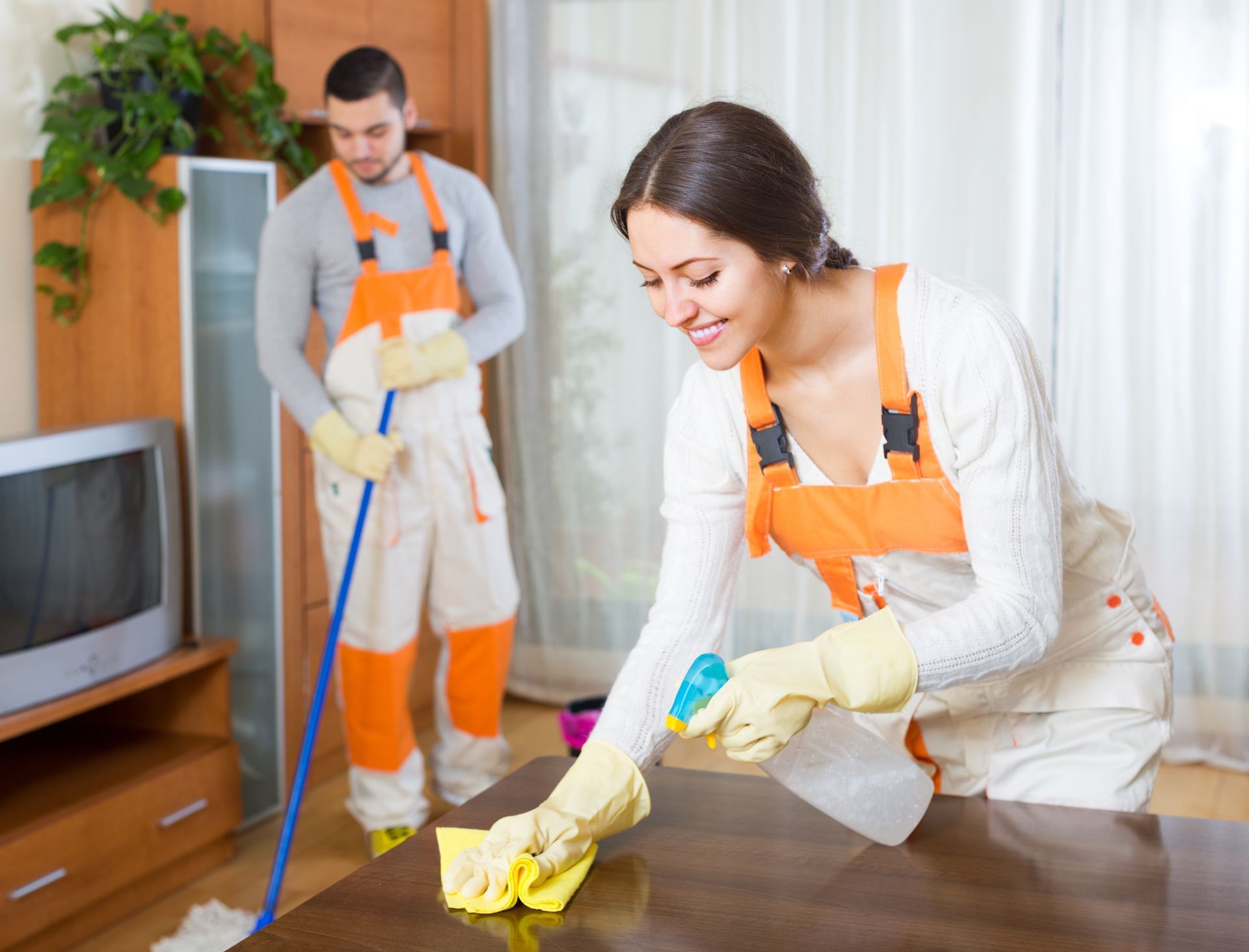 Cleaners in protective gloves wiping surfaces and mopping floors inside a furnished living room