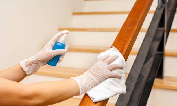 Cleaning wooden stair handrail with disinfectant spray and cloth, home staircase sanitation service.