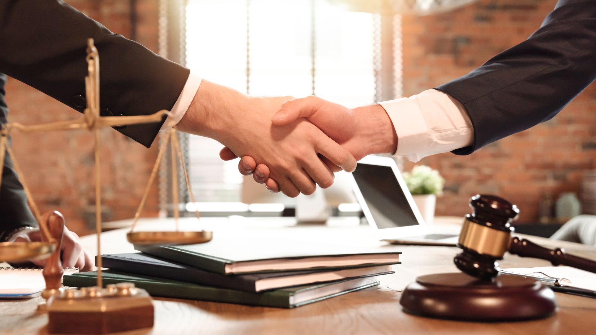 Two people in suits shaking hands in a law office, with a gavel and scales of justice on the table.