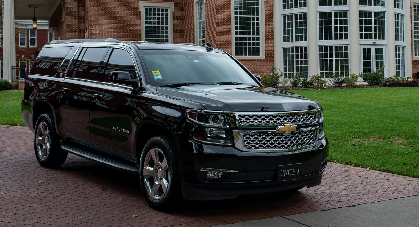 A black Chevrolet Suburban parked on a brick driveway in front of a large brick building with large windows.