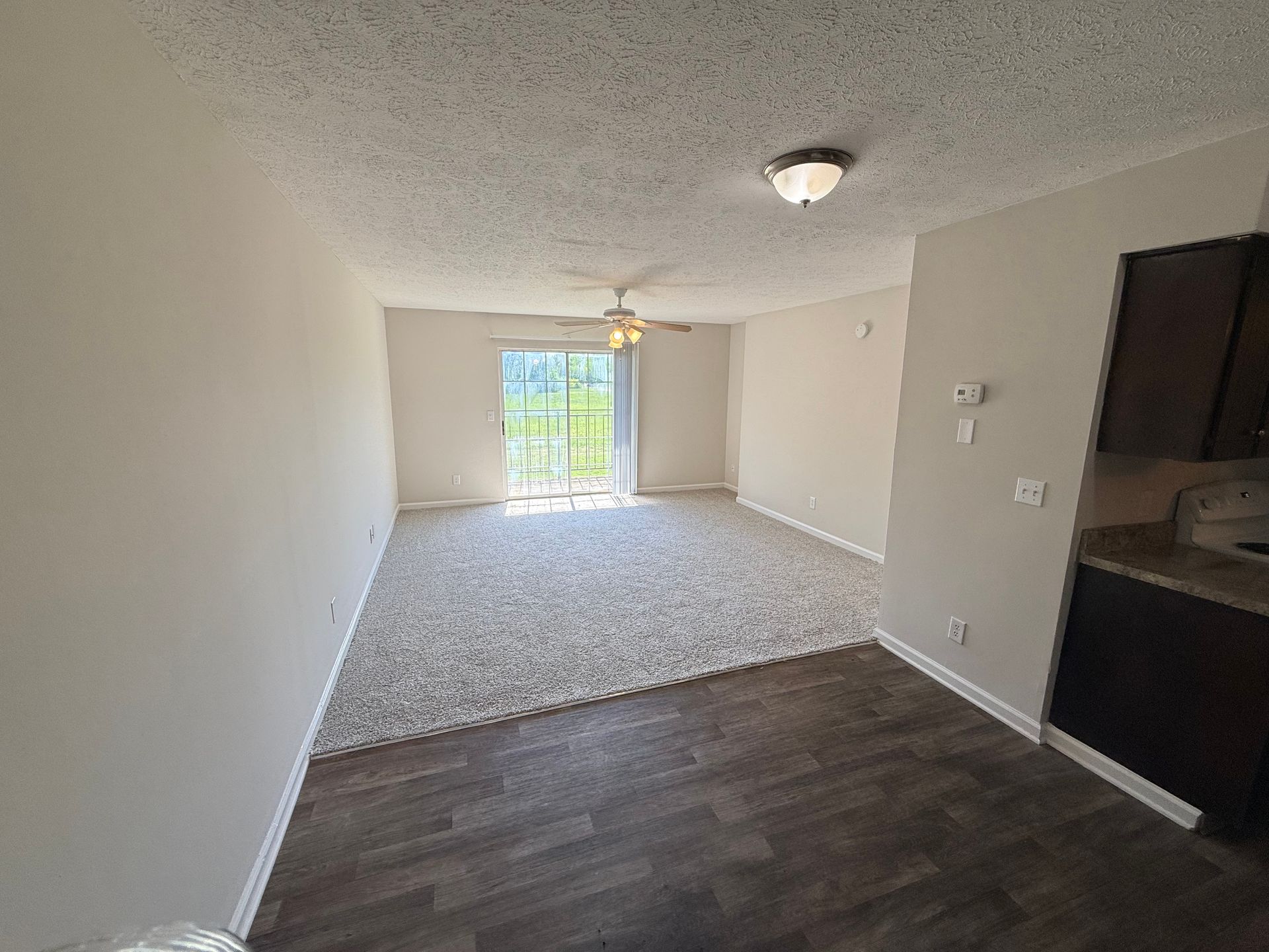 Empty apartment living area with dark wood floor, white walls, carpeted section, and sliding glass doors to a balcony