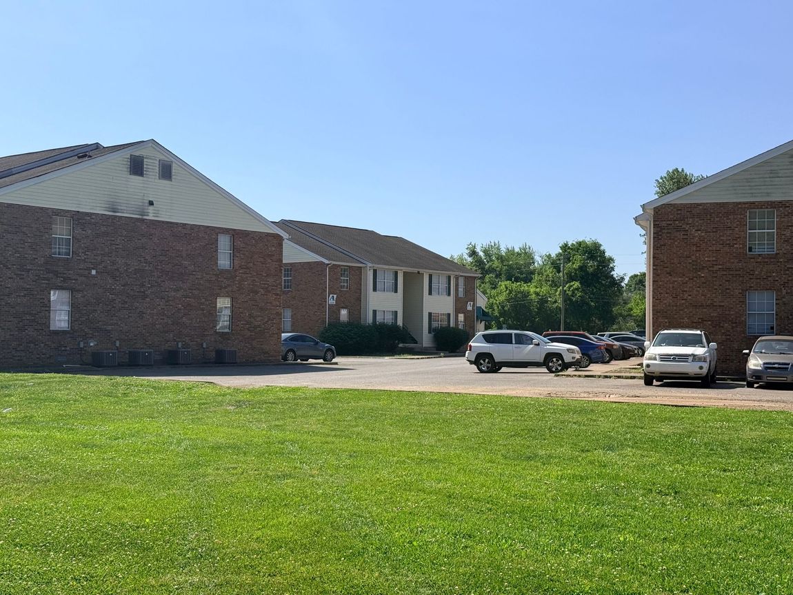 Apartment buildings and parked cars behind a large green grassy field on a clear day
