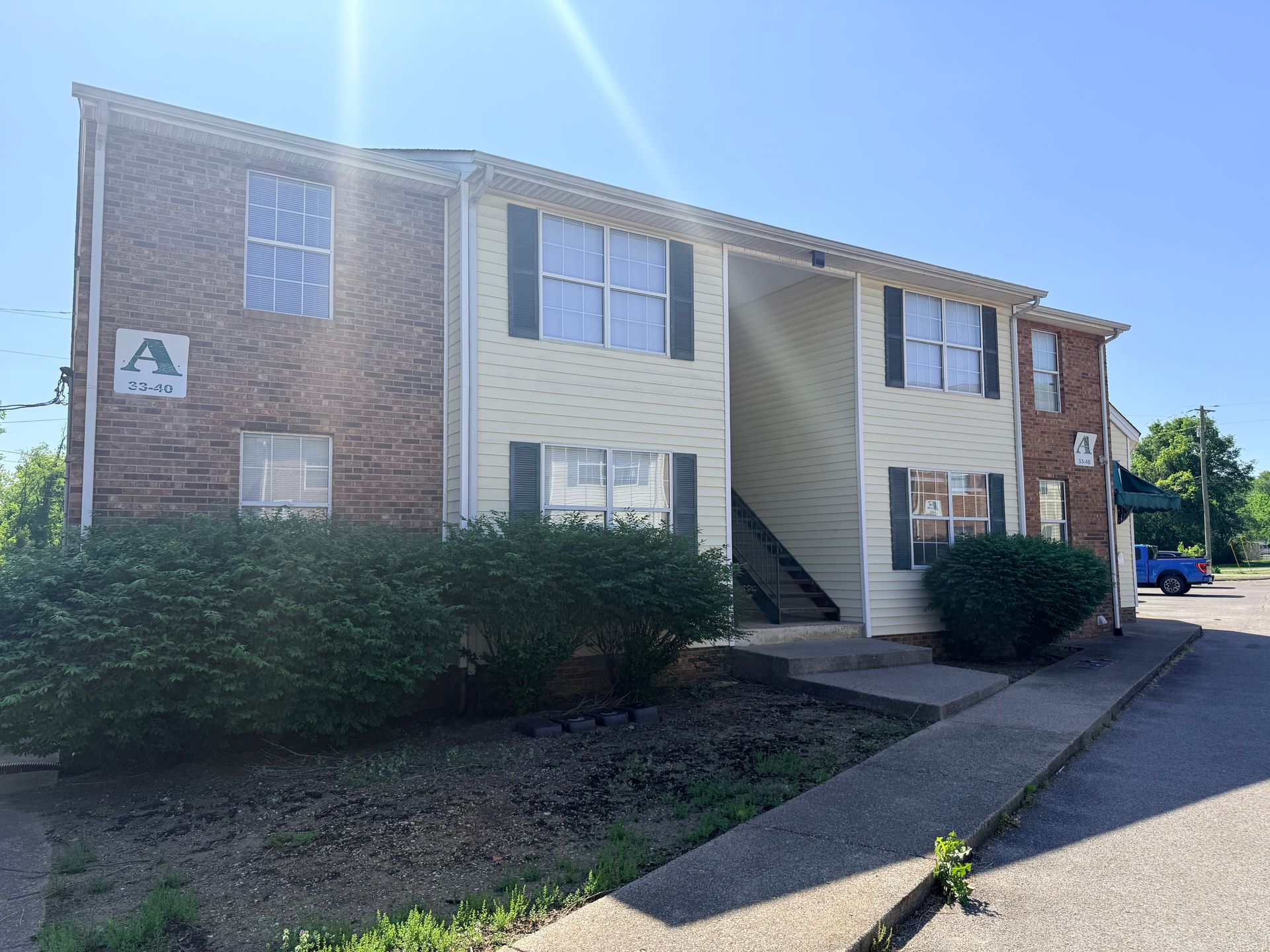 Apartment building with brick and beige siding, central stairwell, and bushes along the front walkway.