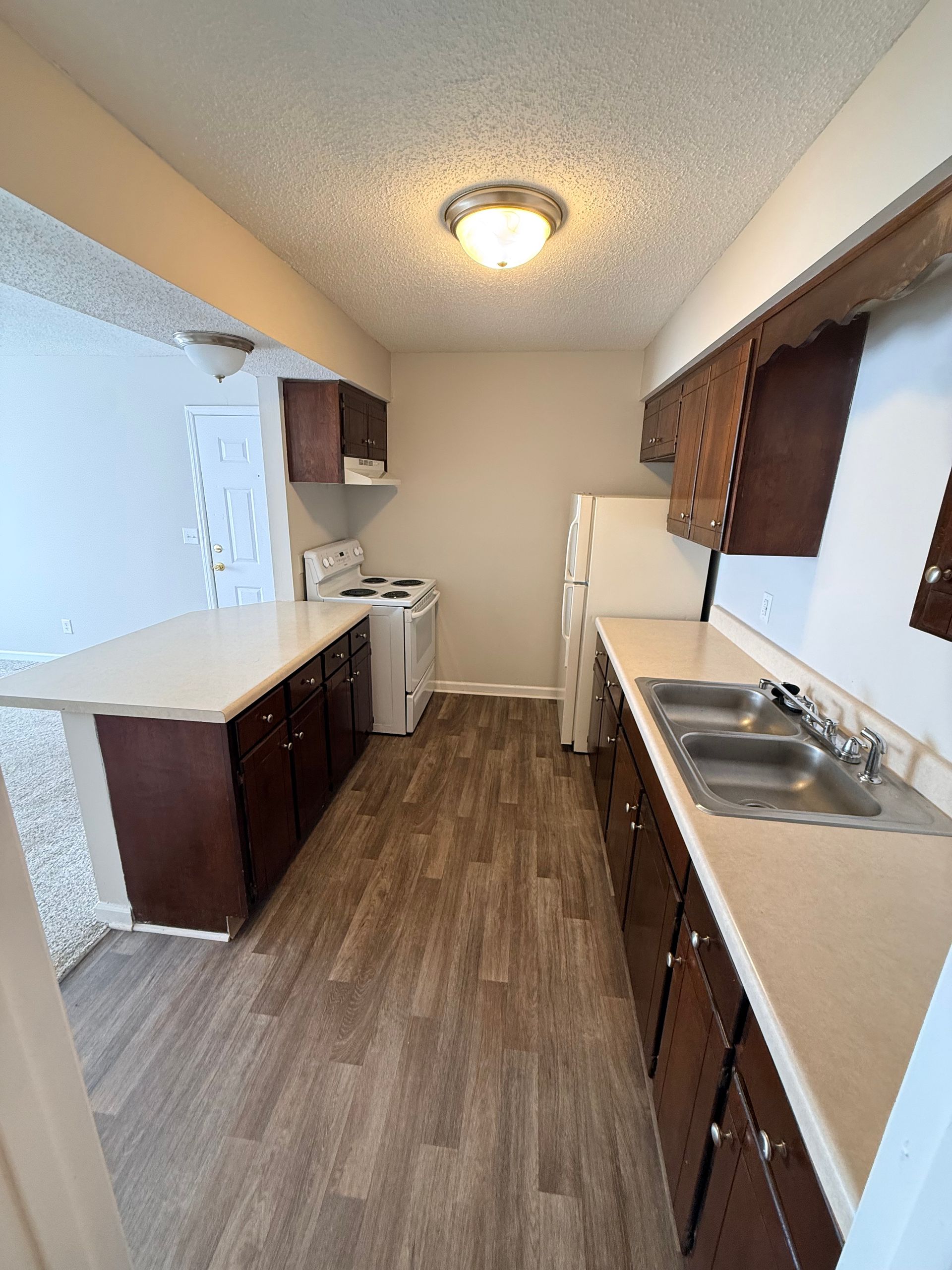Narrow galley kitchen with wood cabinets, white countertops, sink, stove, and vinyl plank flooring.