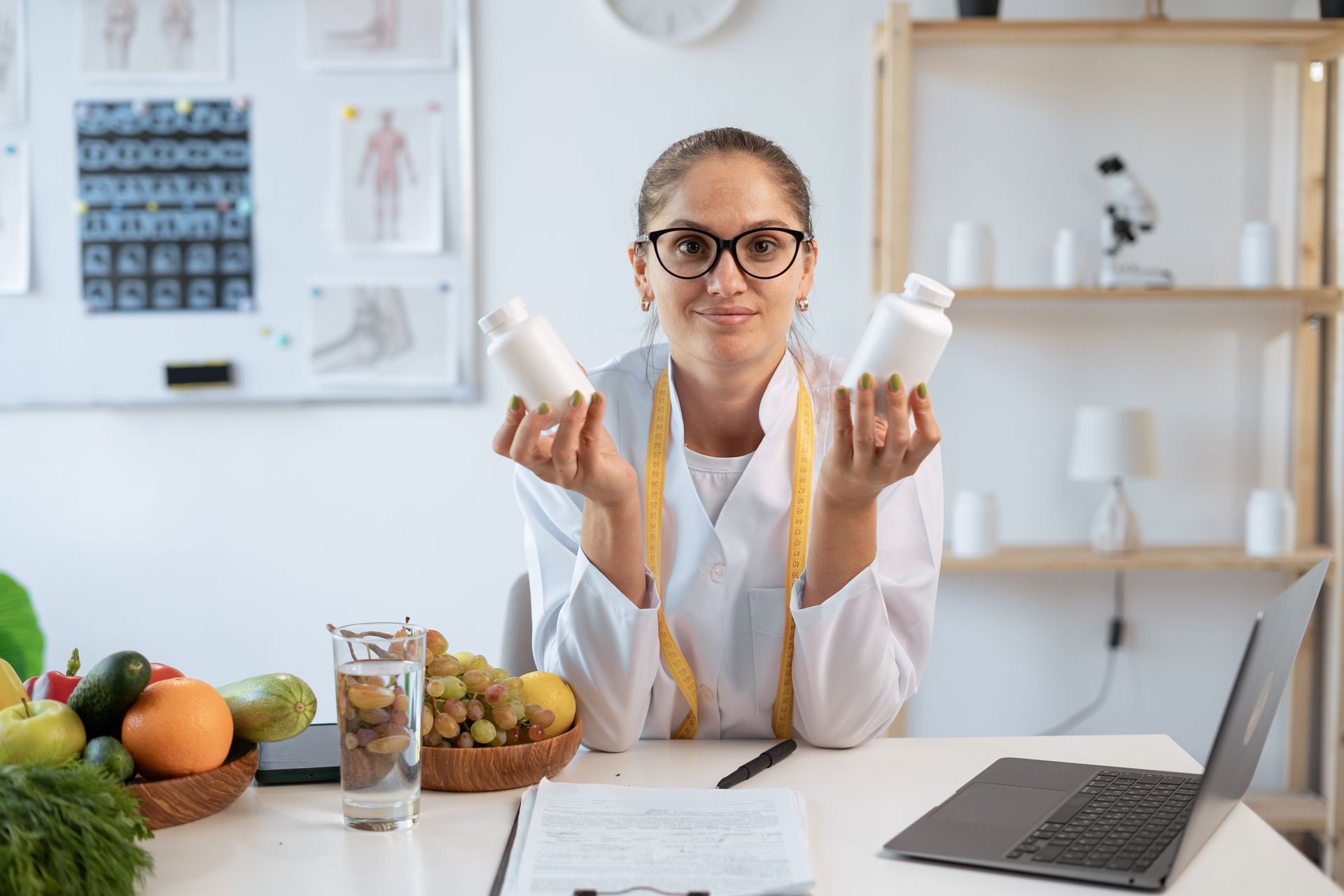 Nutritionist holding supplement bottles, seated at a desk with fruits, water, and laptop.