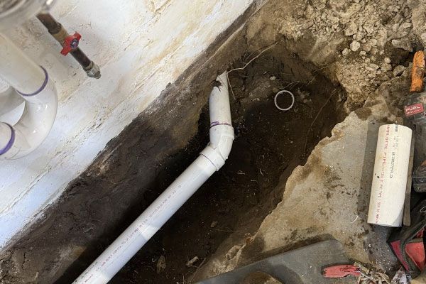 Exposed PVC drain pipes beside a dug-up floor trench with tools and debris in a utility area