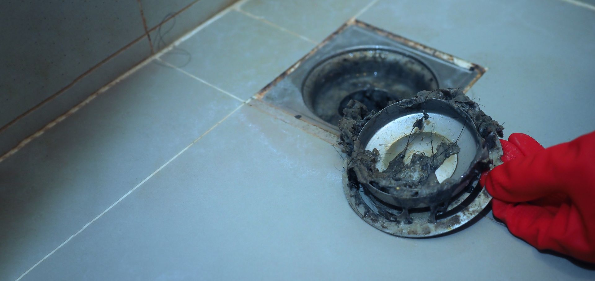 Red-gloved hand holding a dirty drain cover over a tiled floor drain.