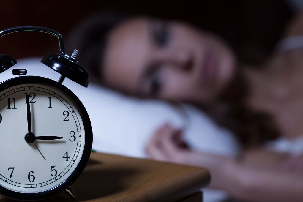 Sveglia nera in primo piano con una persona sfocata sdraiata a letto sullo sfondo, a suggerire l'ora del risveglio mattutino.