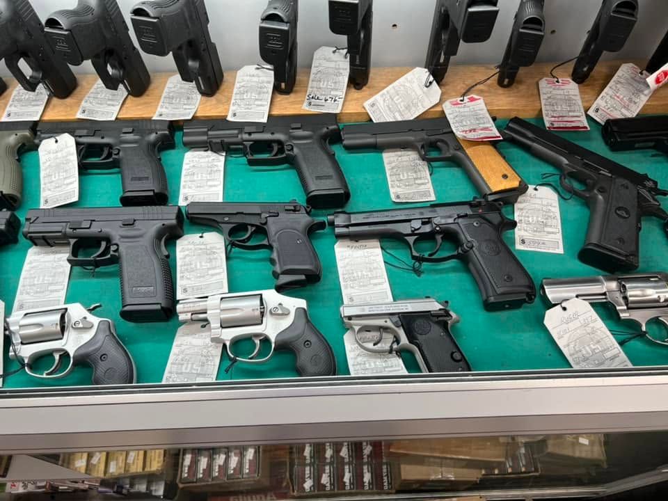 Assorted handguns displayed in a glass case on green lining with price tags and magazines.