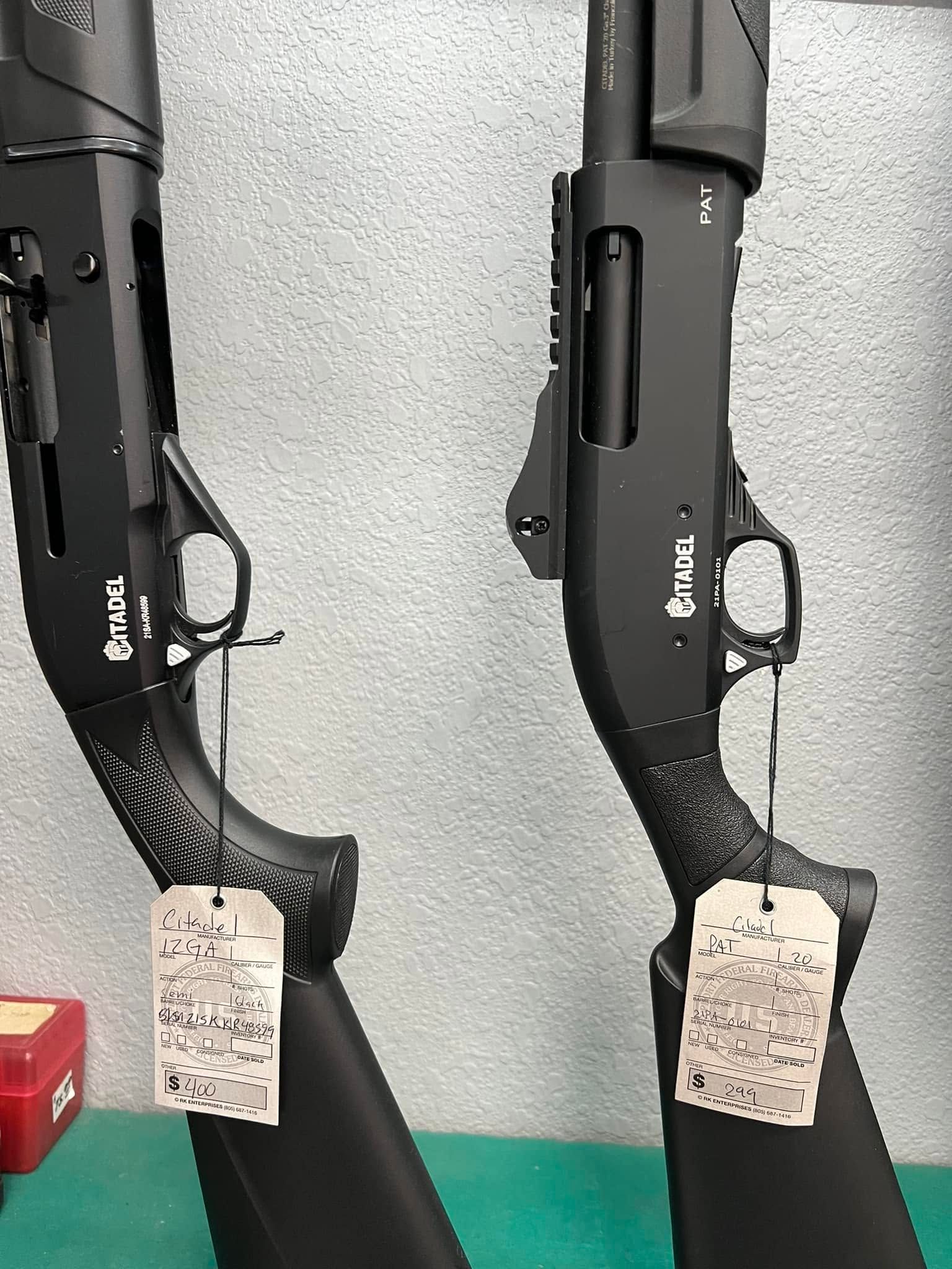Two black shotguns displayed upright on stands with hanging tags against a gray wall