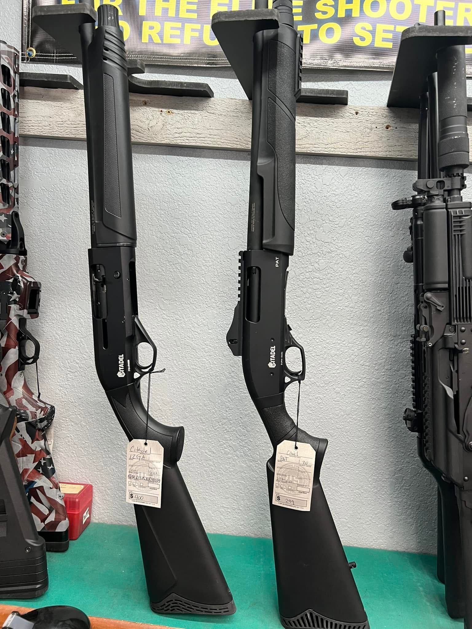 Two black rifles displayed upright on a wall in a gun shop, with price tags near the stocks