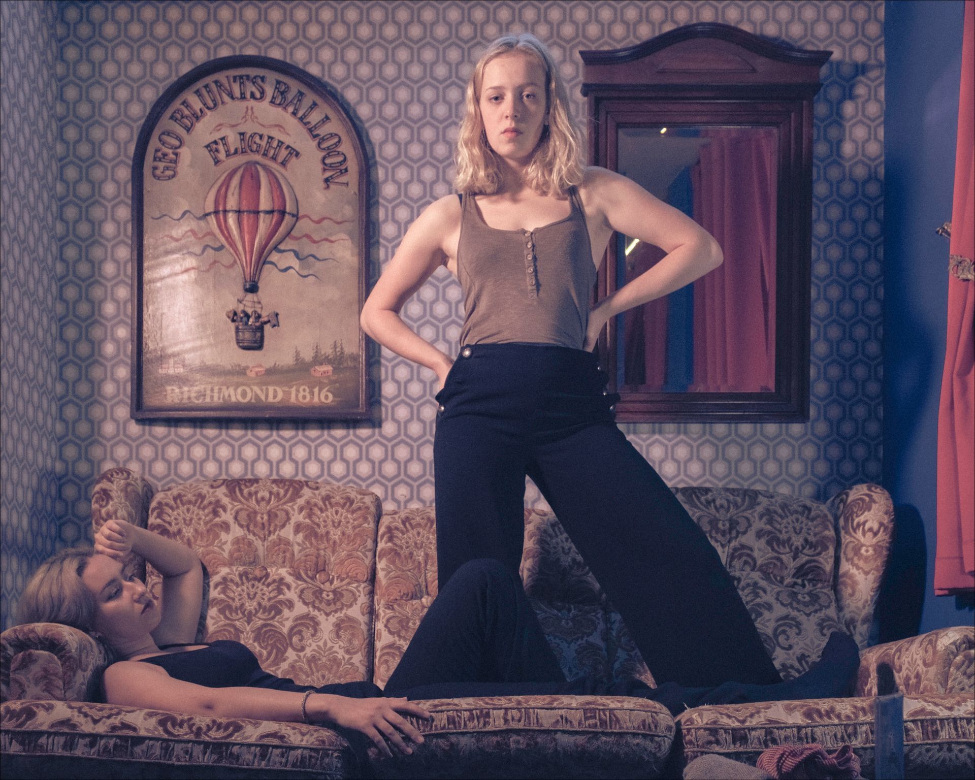 Two women in a room. One stands, hands on hips. The other reclines on a patterned sofa.  A poster and mirror are on the wall.