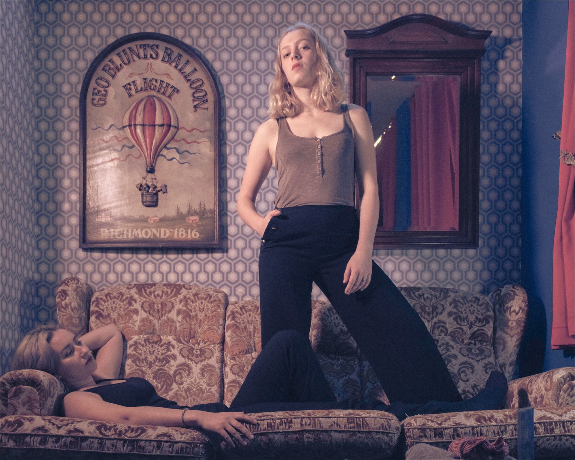 Two women pose on a patterned sofa. One stands, the other reclines. A hot air balloon poster hangs on the wall.