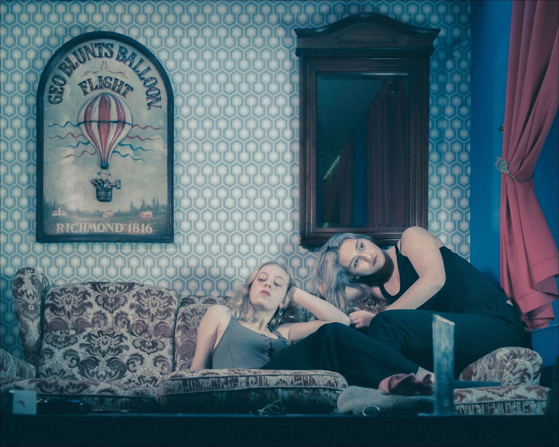 Two people on a patterned couch, one lounging, other leaning. Wallpaper, mirror, and a hot air balloon poster