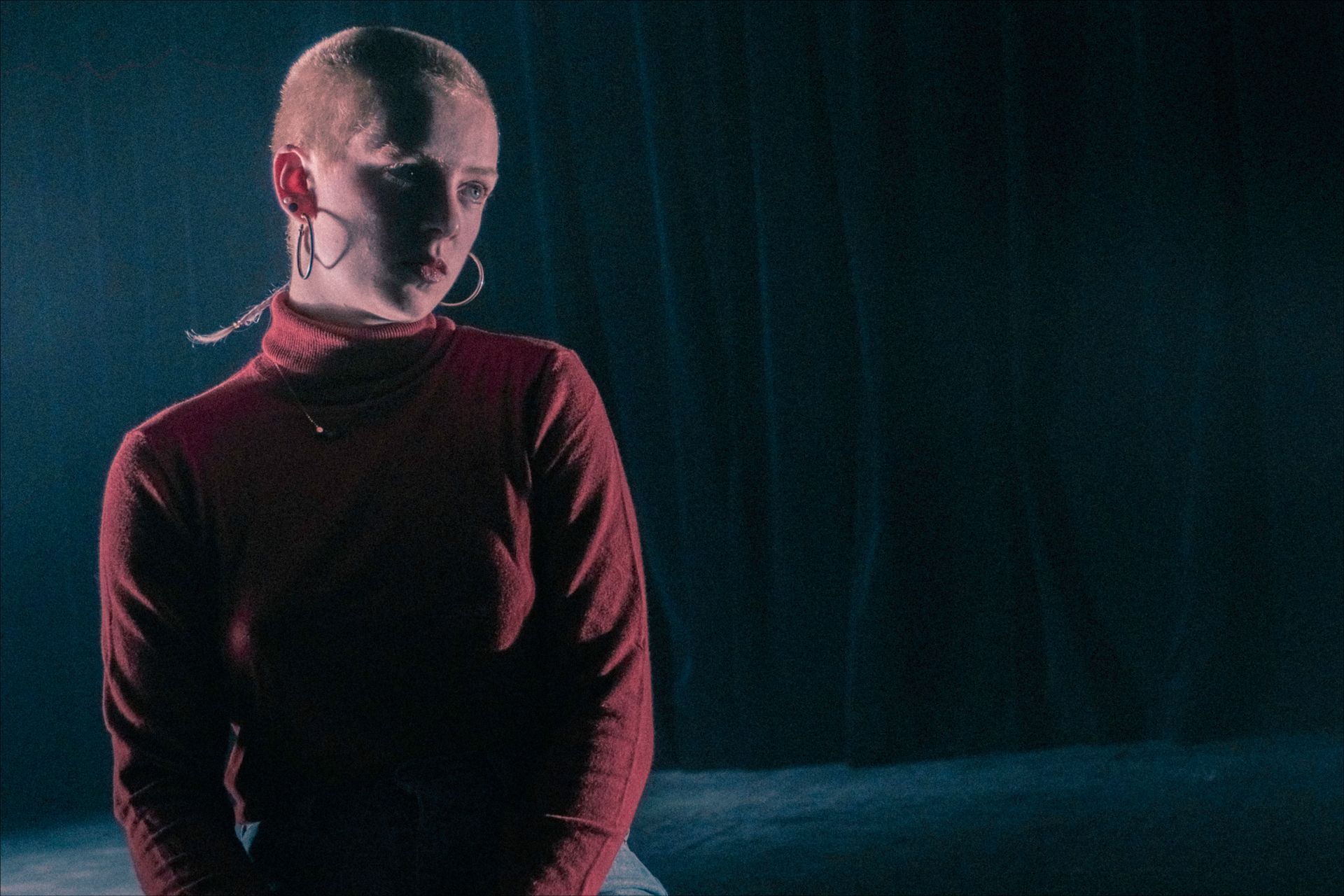 Woman with shaved head, wearing a red turtleneck and hoop earrings, looking down in a dimly lit space.