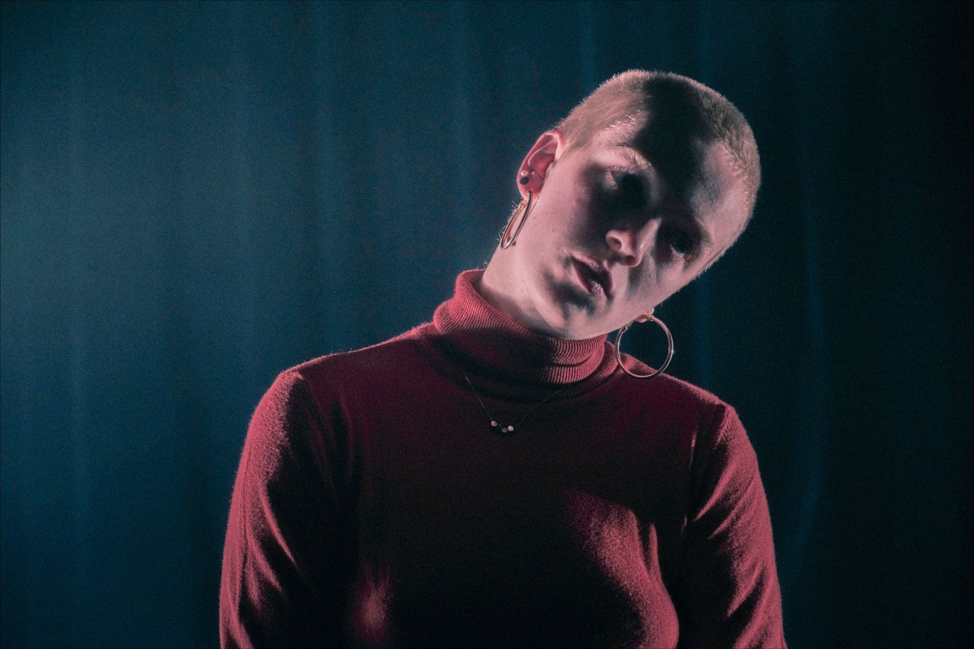 Woman with shaved head in red turtleneck, head tilted, against a dark blue background.