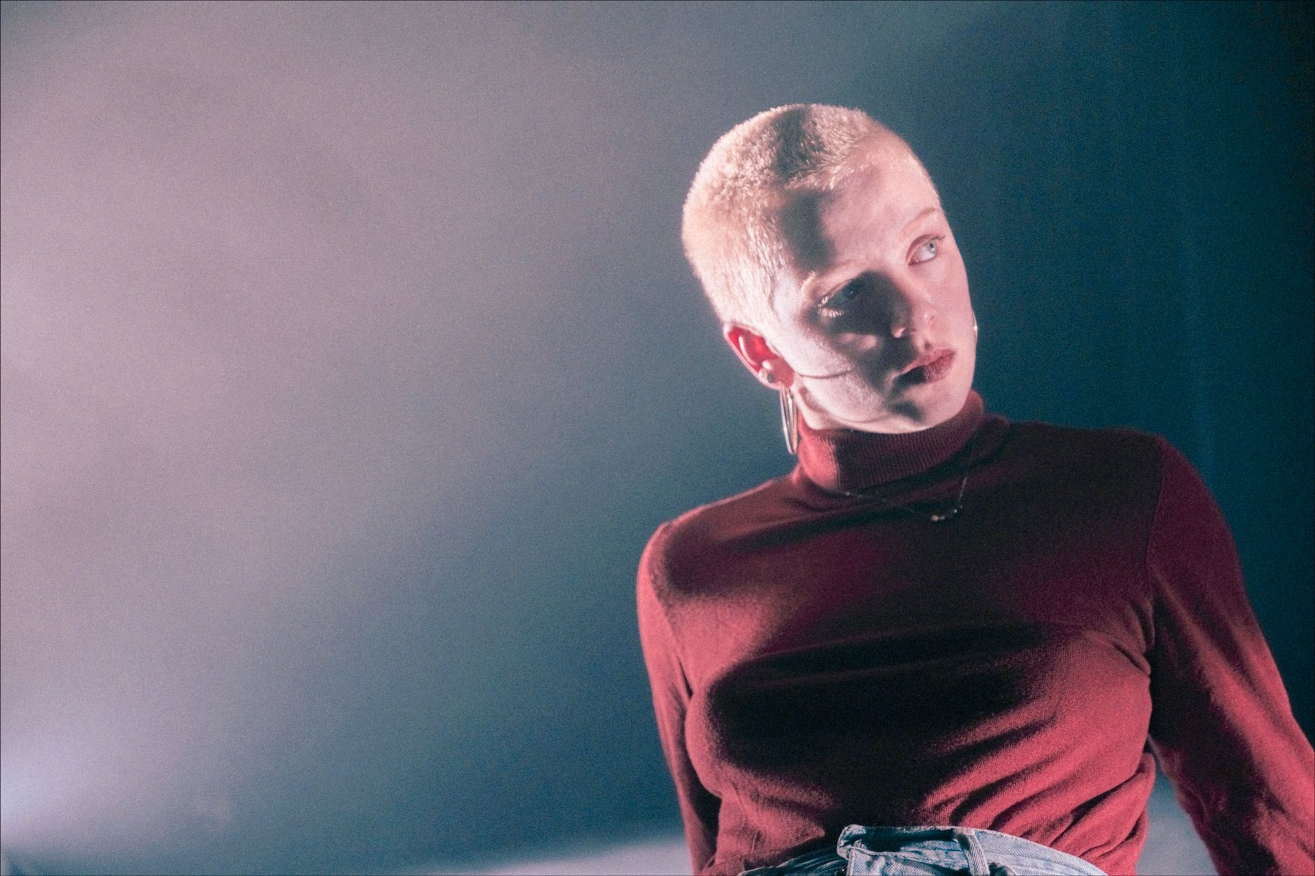 Person with shaved head in a red turtleneck, looking slightly up and to the right, against a smoky background.
