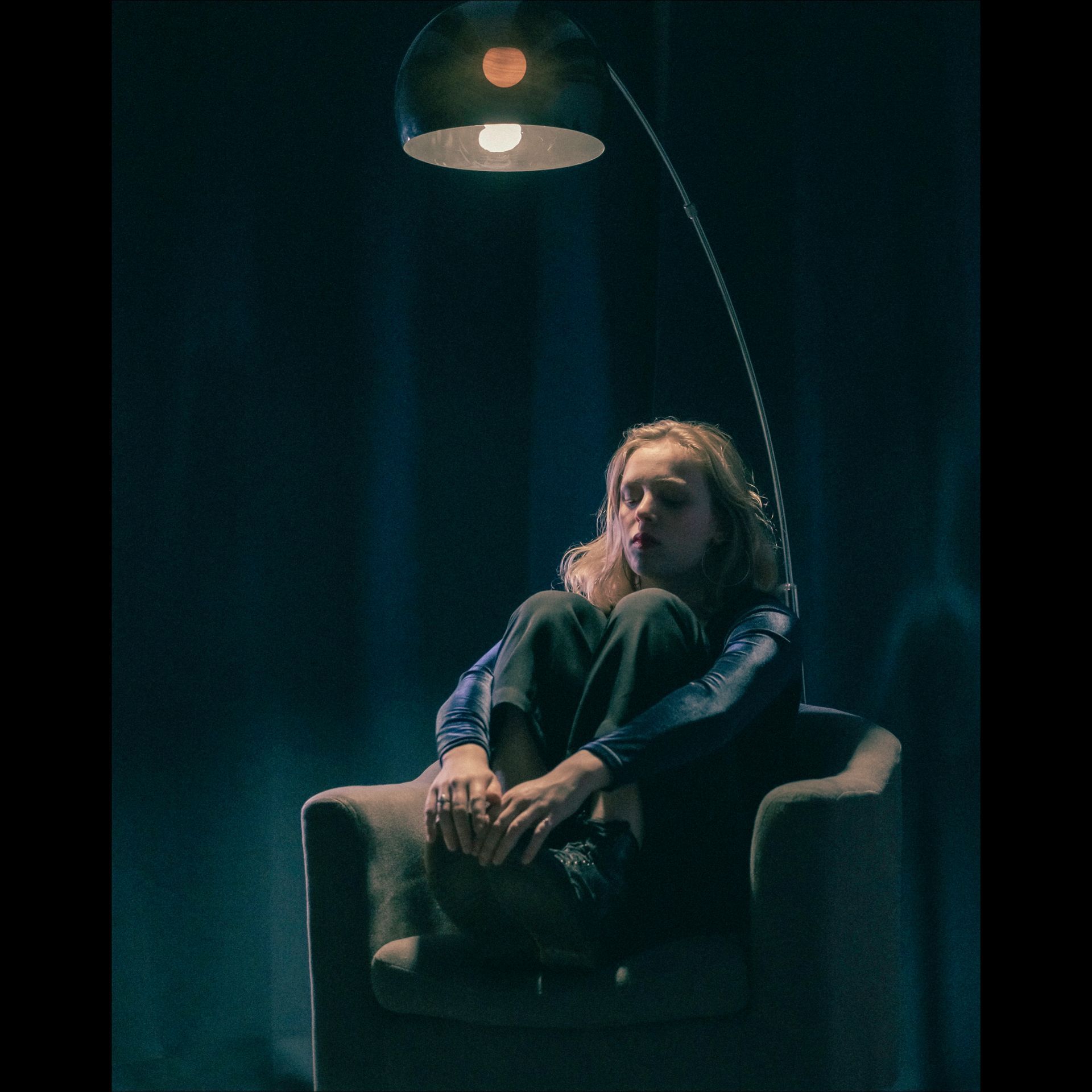 Person sitting in chair under a lamp, looking thoughtful. Dark setting.