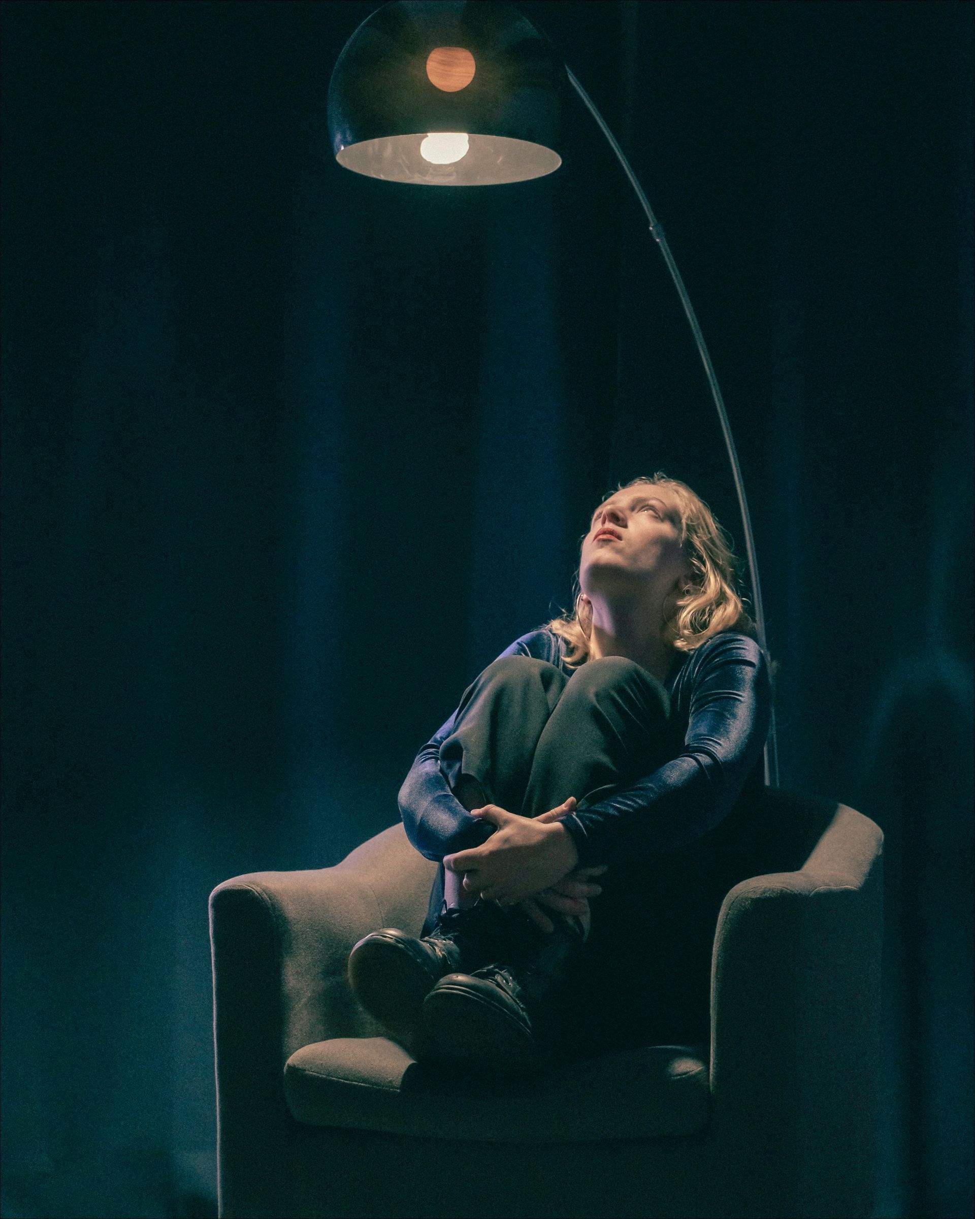 Woman in a chair, looking up at a lit lamp, in a dim room.