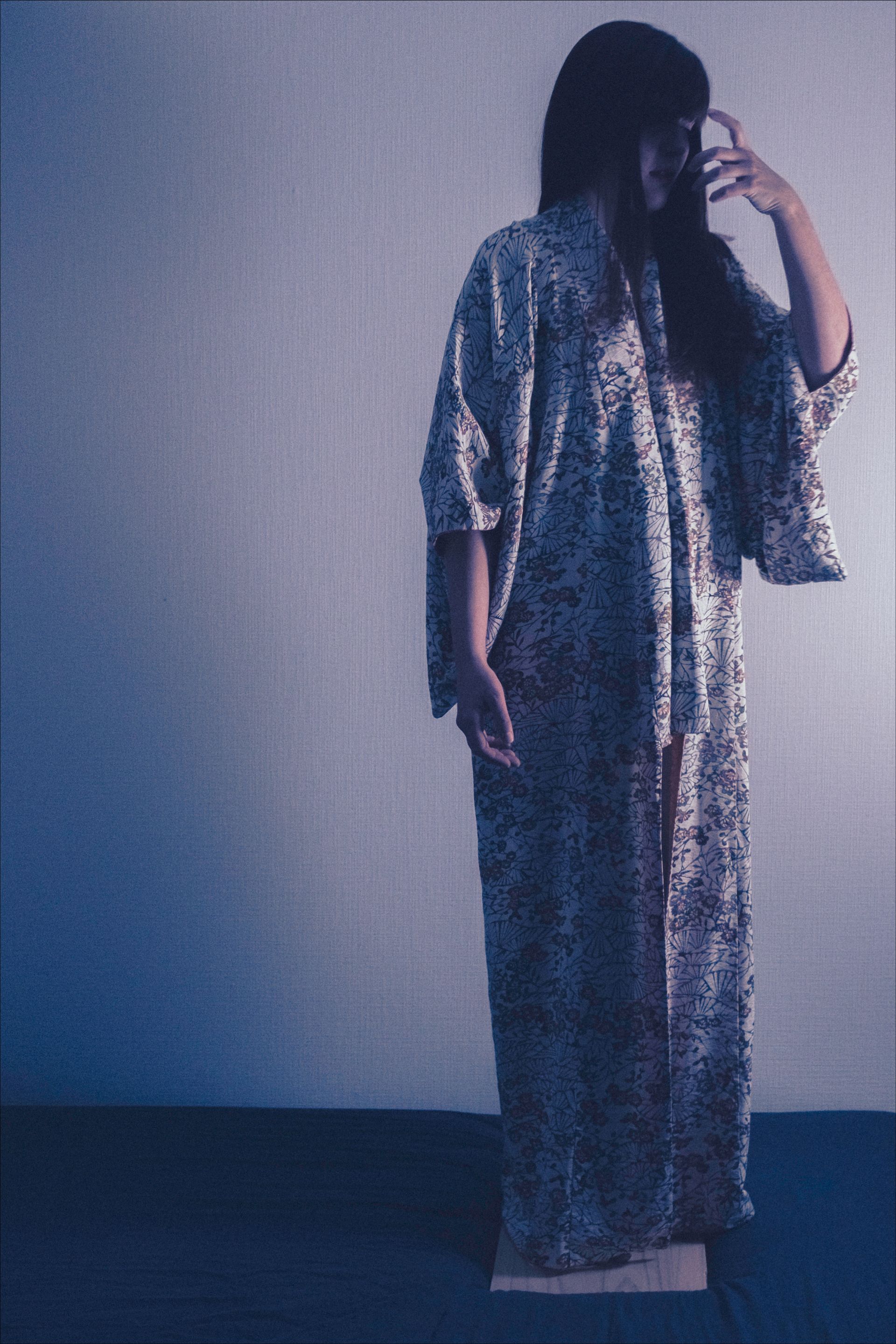 Person in long patterned robe, standing with hand to head, against a blue-lit wall.
