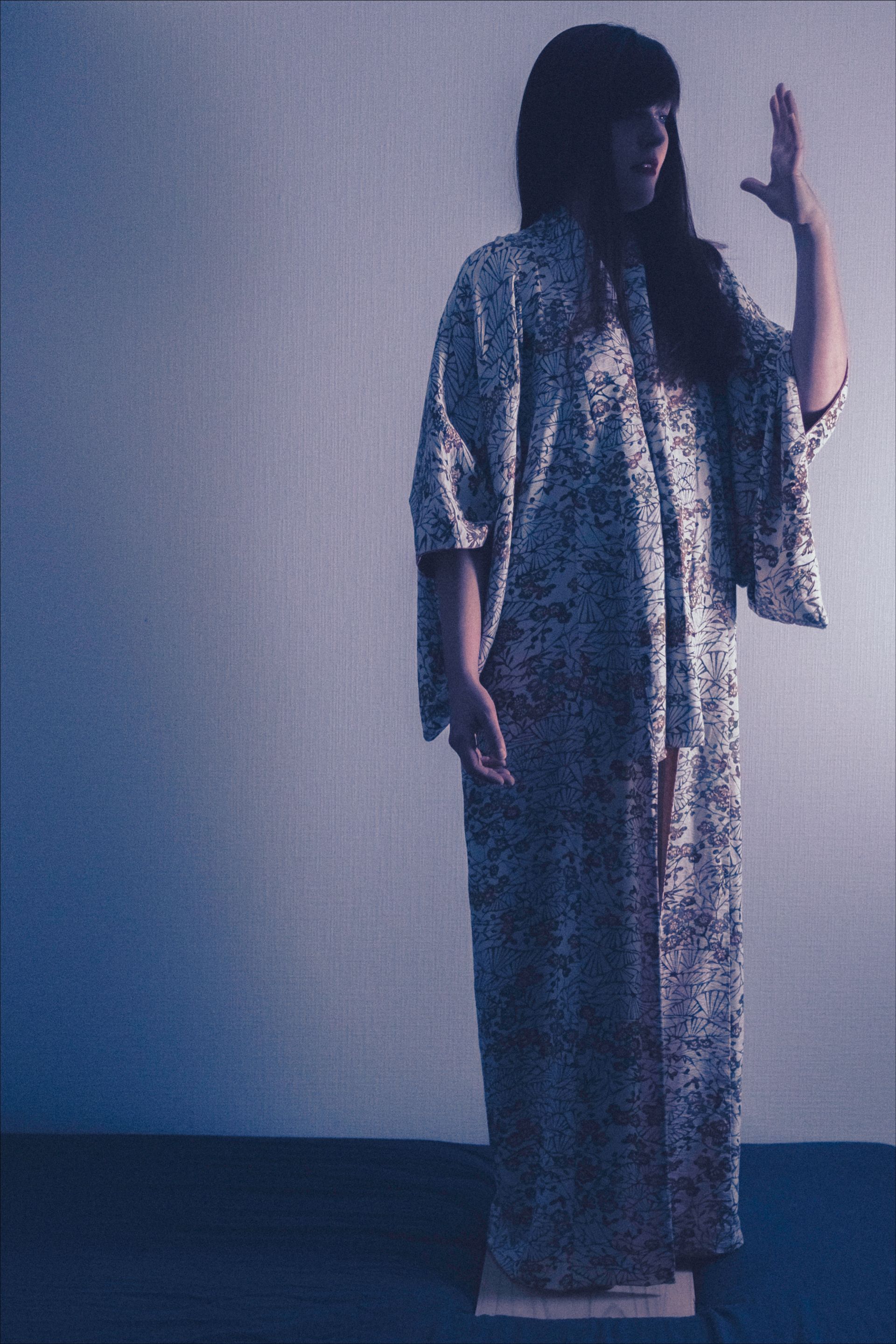 Woman in floral robe, arm raised, standing. Soft lighting.