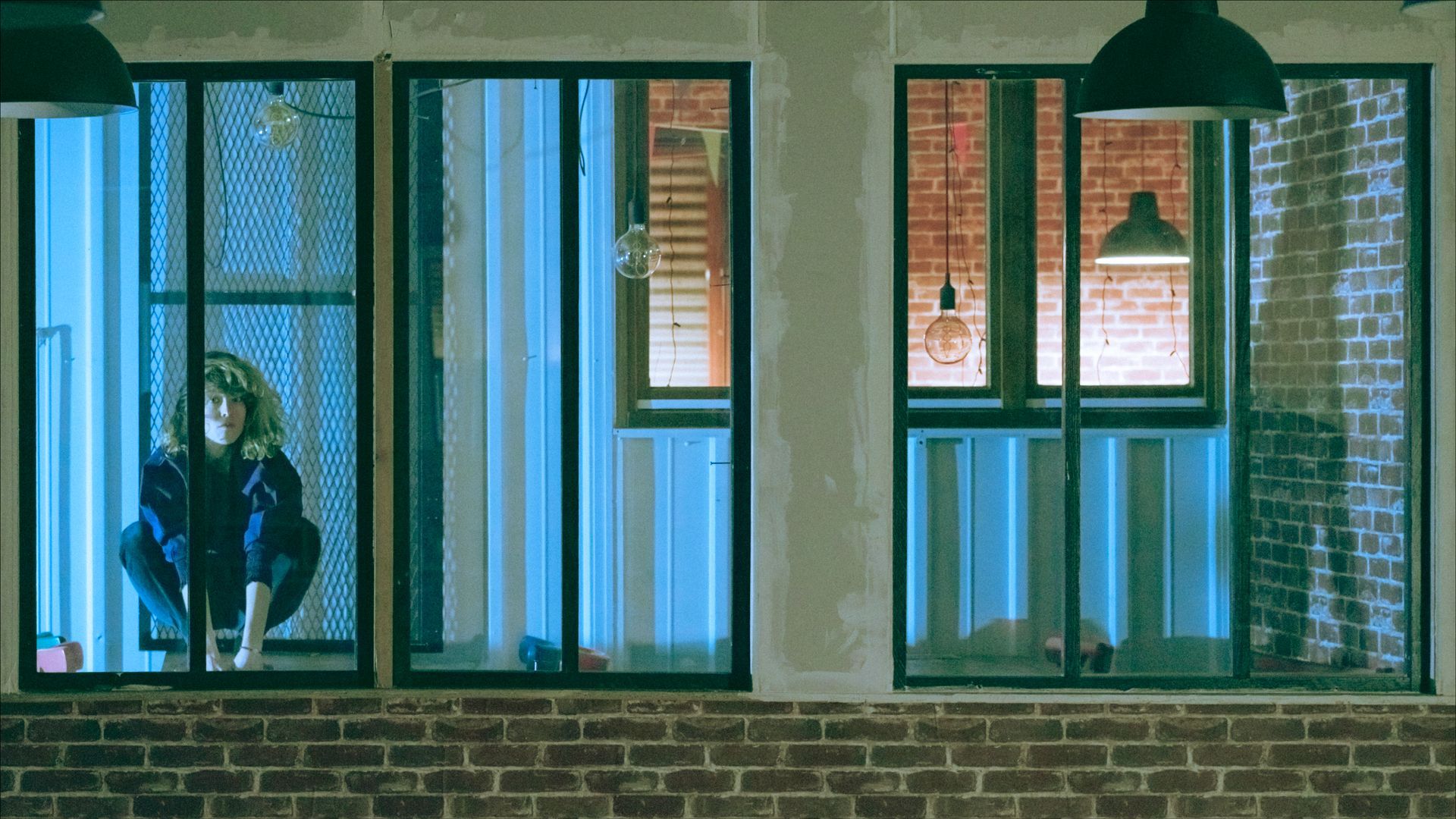 Woman squatting in a blue-lit window, looking down. Brick exterior with industrial-style windows.