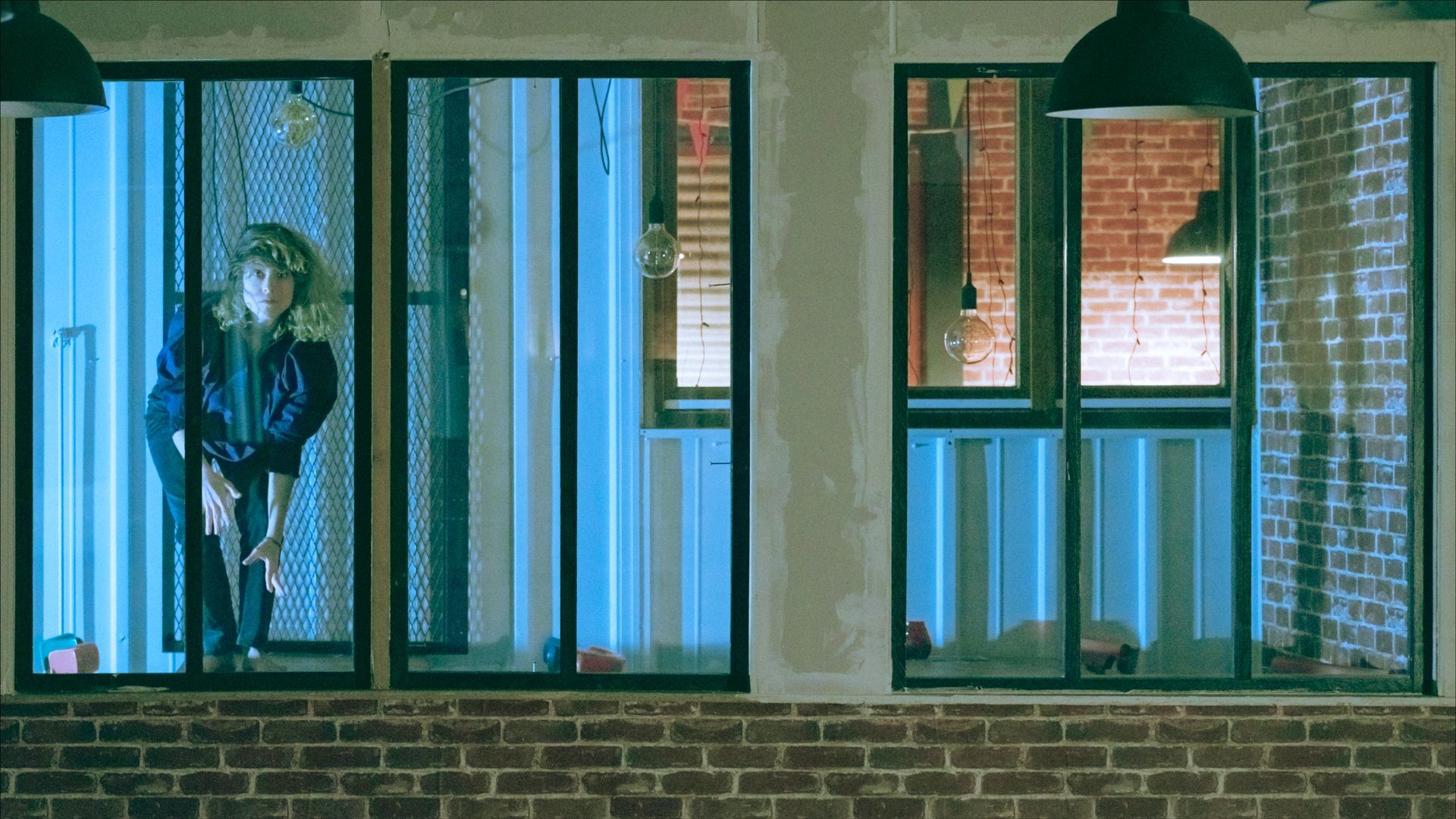 Woman in blue jacket leans against a window with bars, in a building with brick walls.