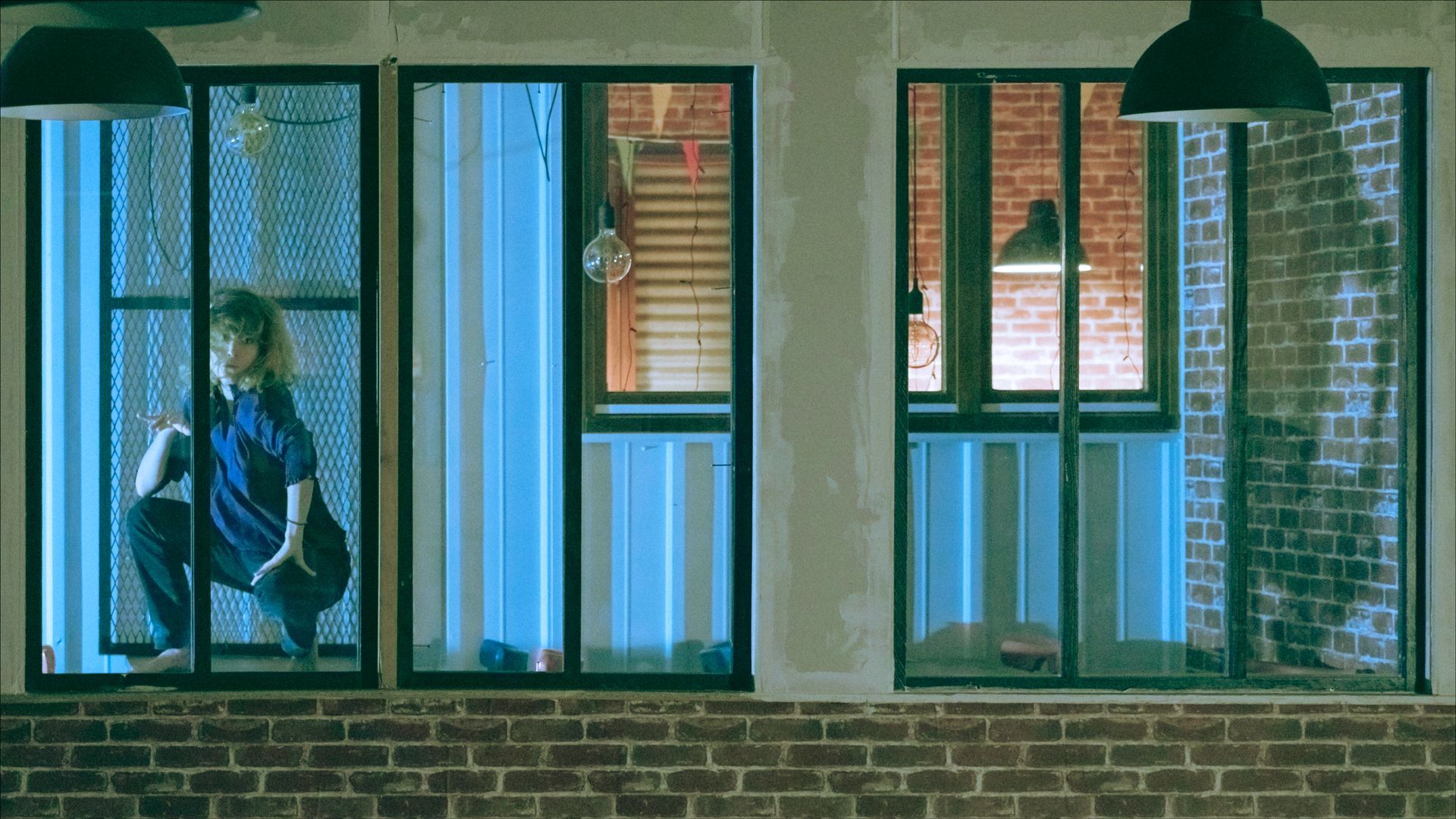 Woman in blue outfit squats by a window, looking out. Low angle, brick wall visible.