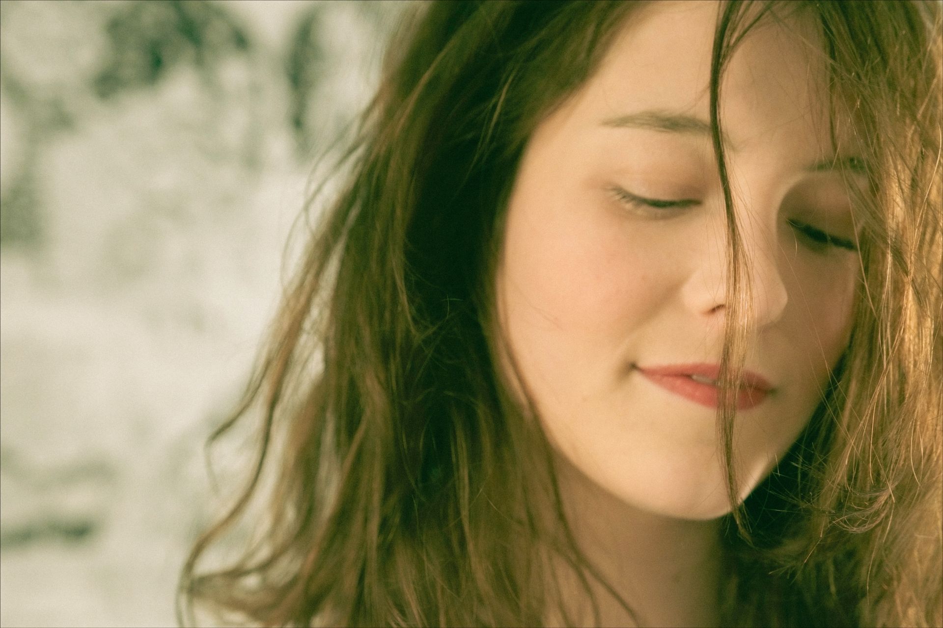 Woman with closed eyes and loose brown hair, slight smile. Soft lighting, neutral background.