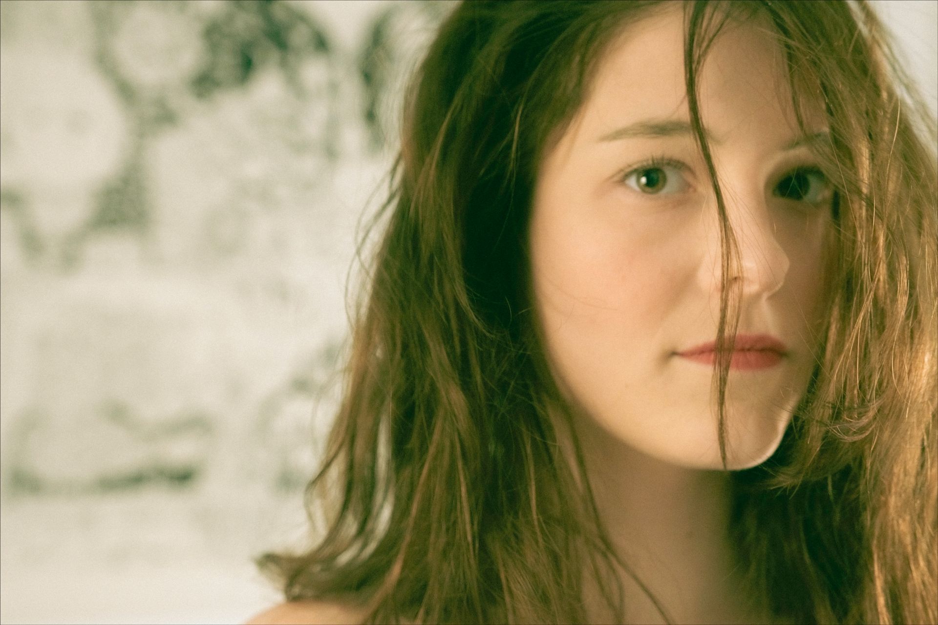 Woman with long brown hair, looking at the camera. A strand of hair hangs over her face. Soft focus background.