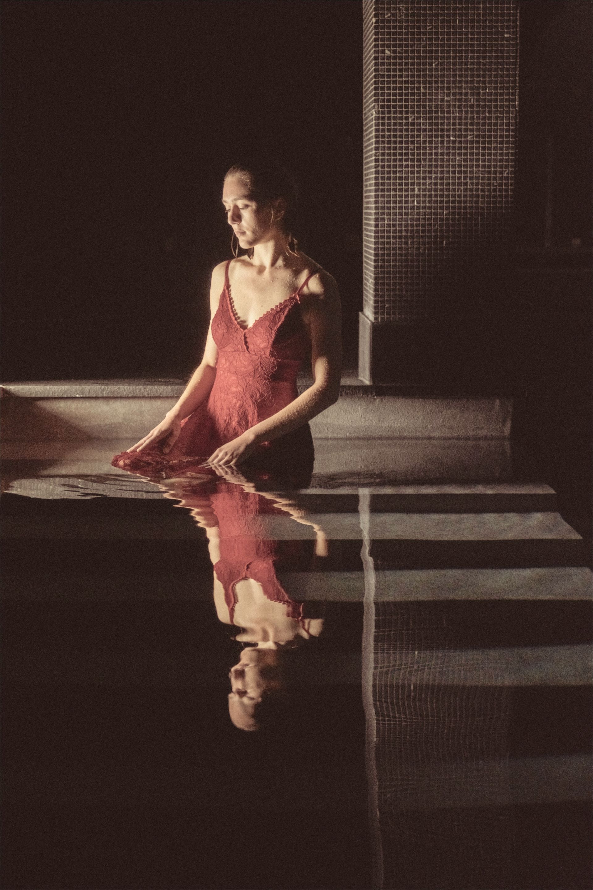 Woman in red dress in water, reflected. Dim lighting, tile wall background.