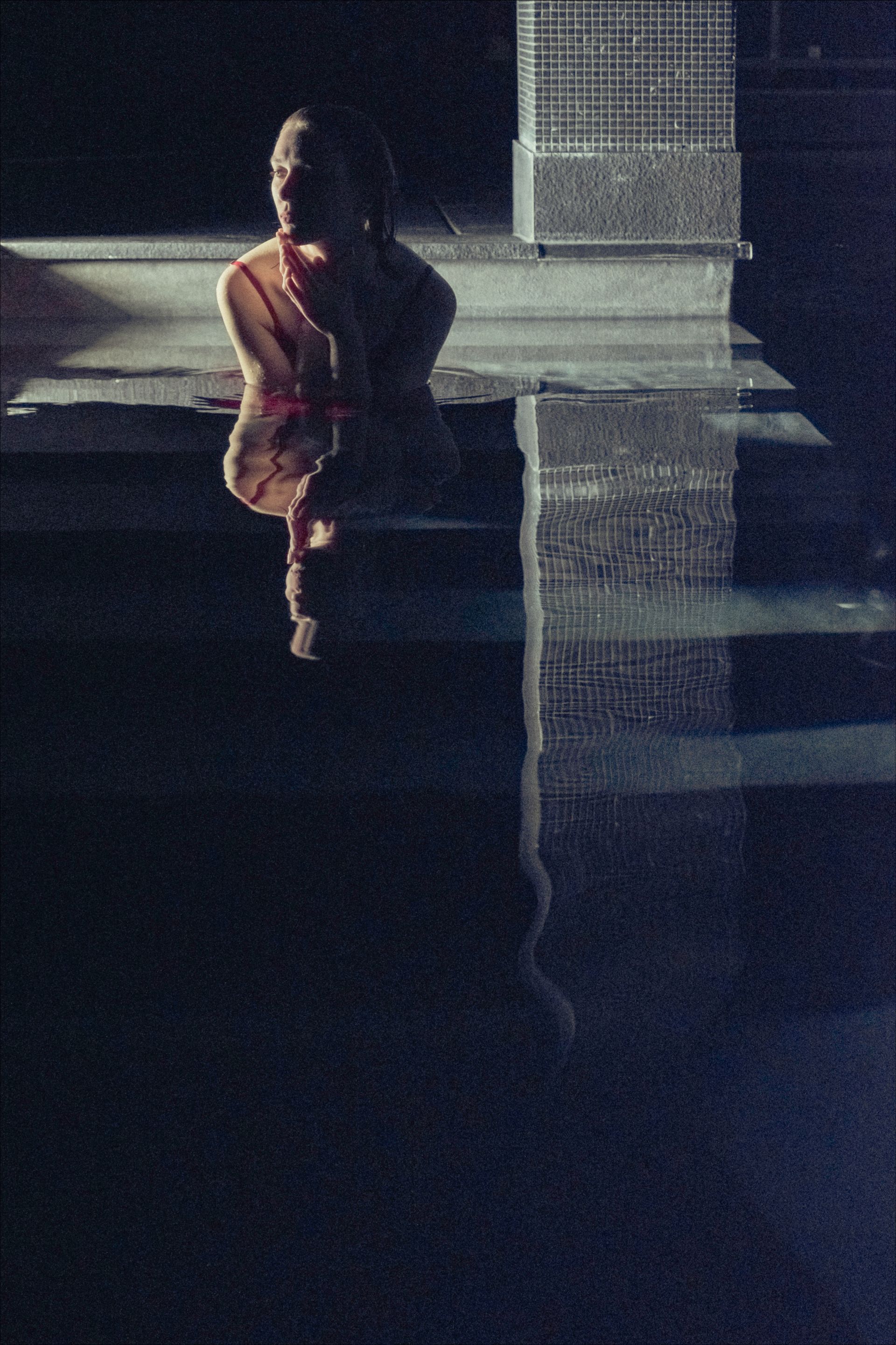 A person sits on a ledge, reflected in dark water. Low light, blurry, watery atmosphere.