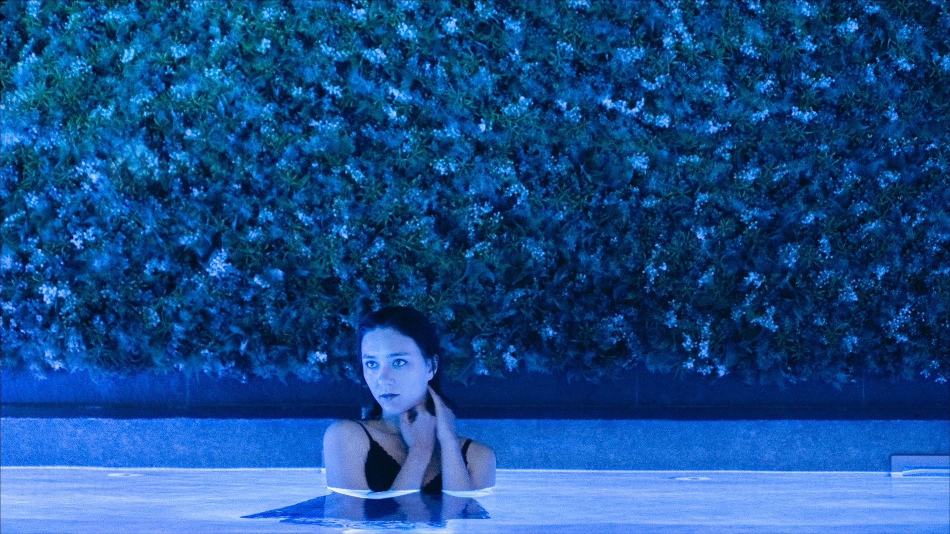 Woman in a black swimsuit in a blue-lit pool, hands near her face, against a dark leafy hedge.