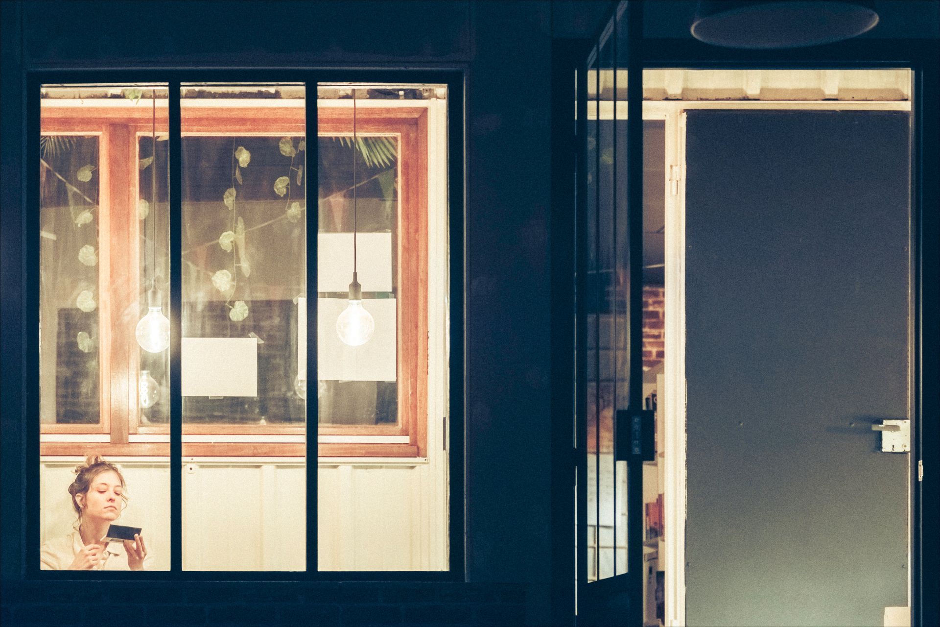 Woman applying makeup at window; interior with open door, ambient light.