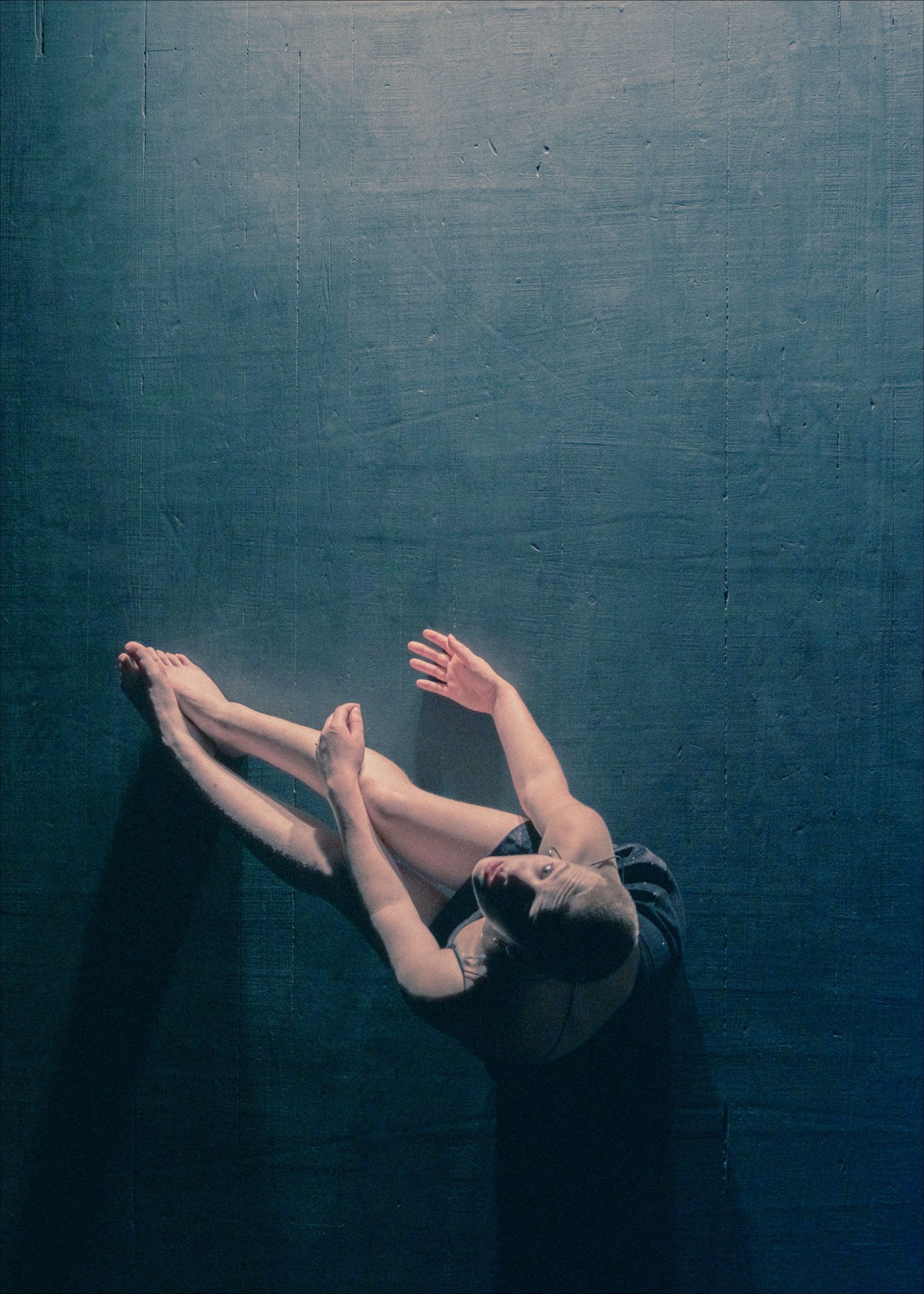 A dancer is on a dark blue stage, stretching with legs extended and arms up.