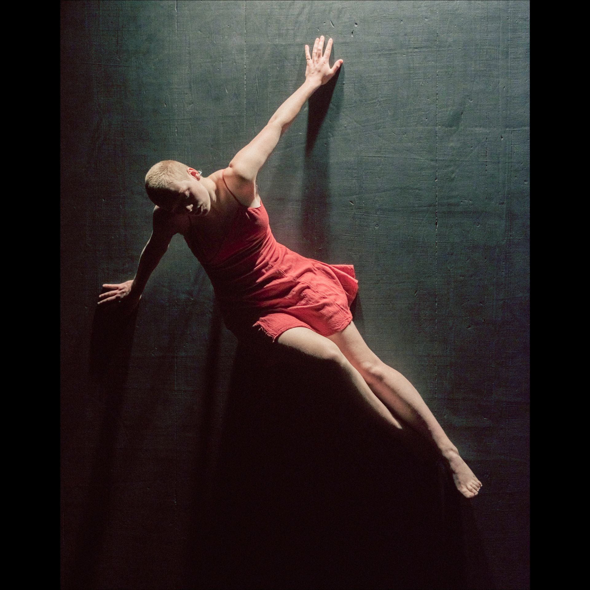 Woman in a red dress leans against a dark wall, right arm extended upwards.