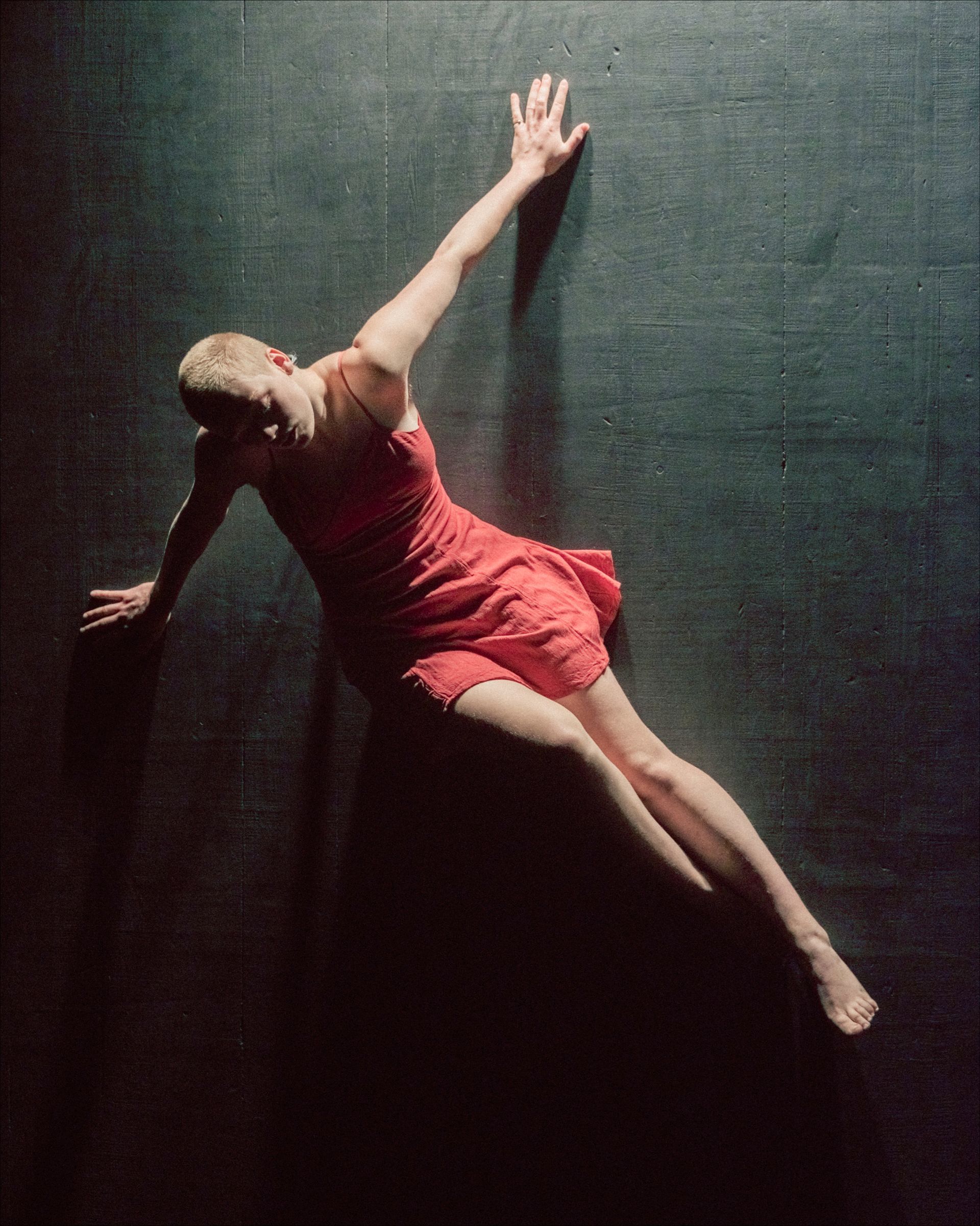 Dancer in red dress leans against a dark wall, right arm extended, leg extended.