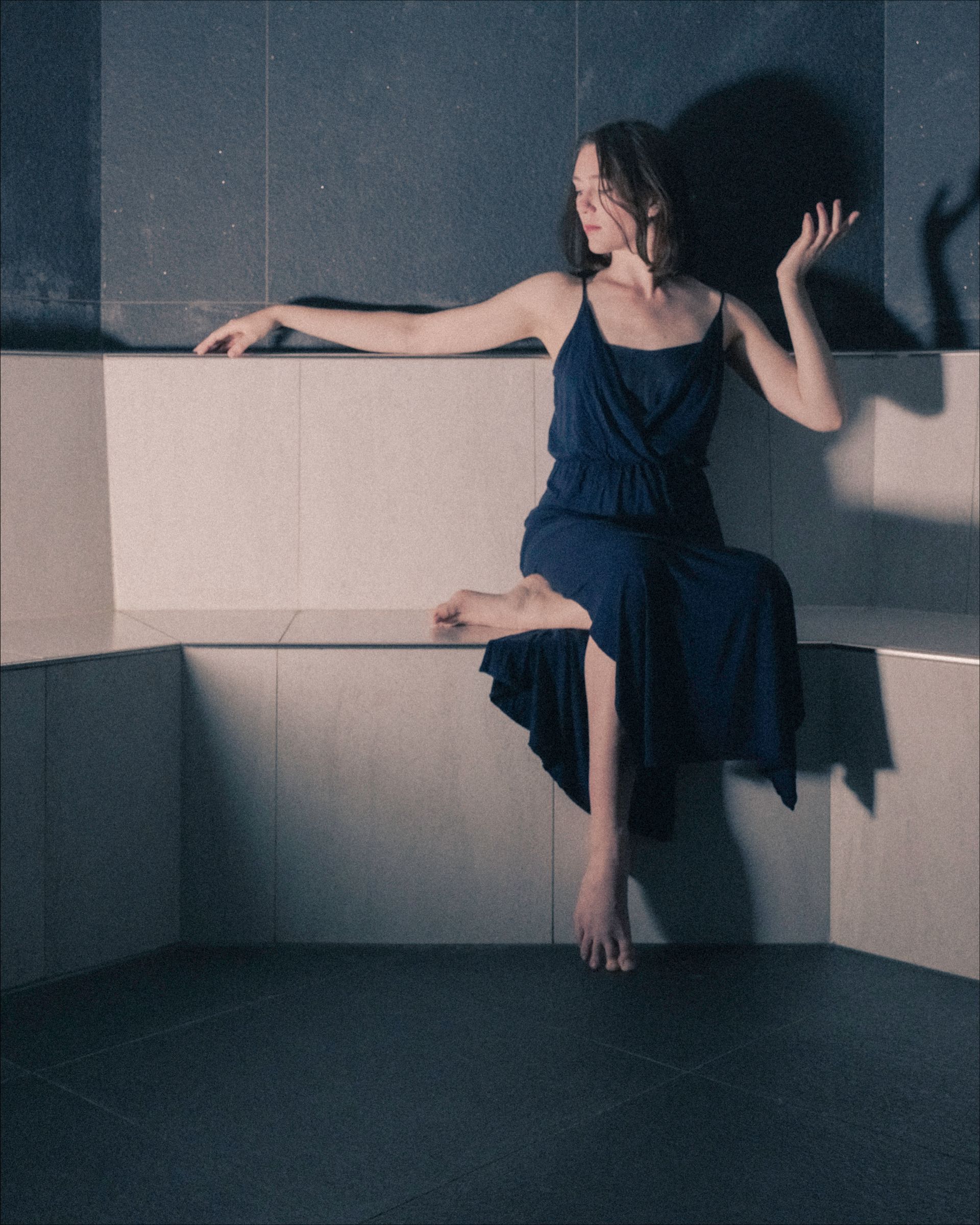 Woman in blue dress sitting on stone bench, one arm outstretched.