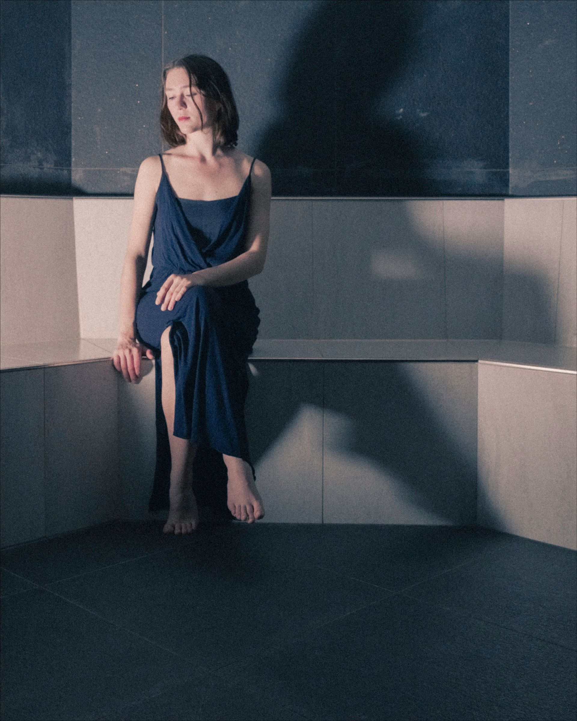 Woman in blue dress sits on a tiled bench, looking away. Dark room, with shadows.