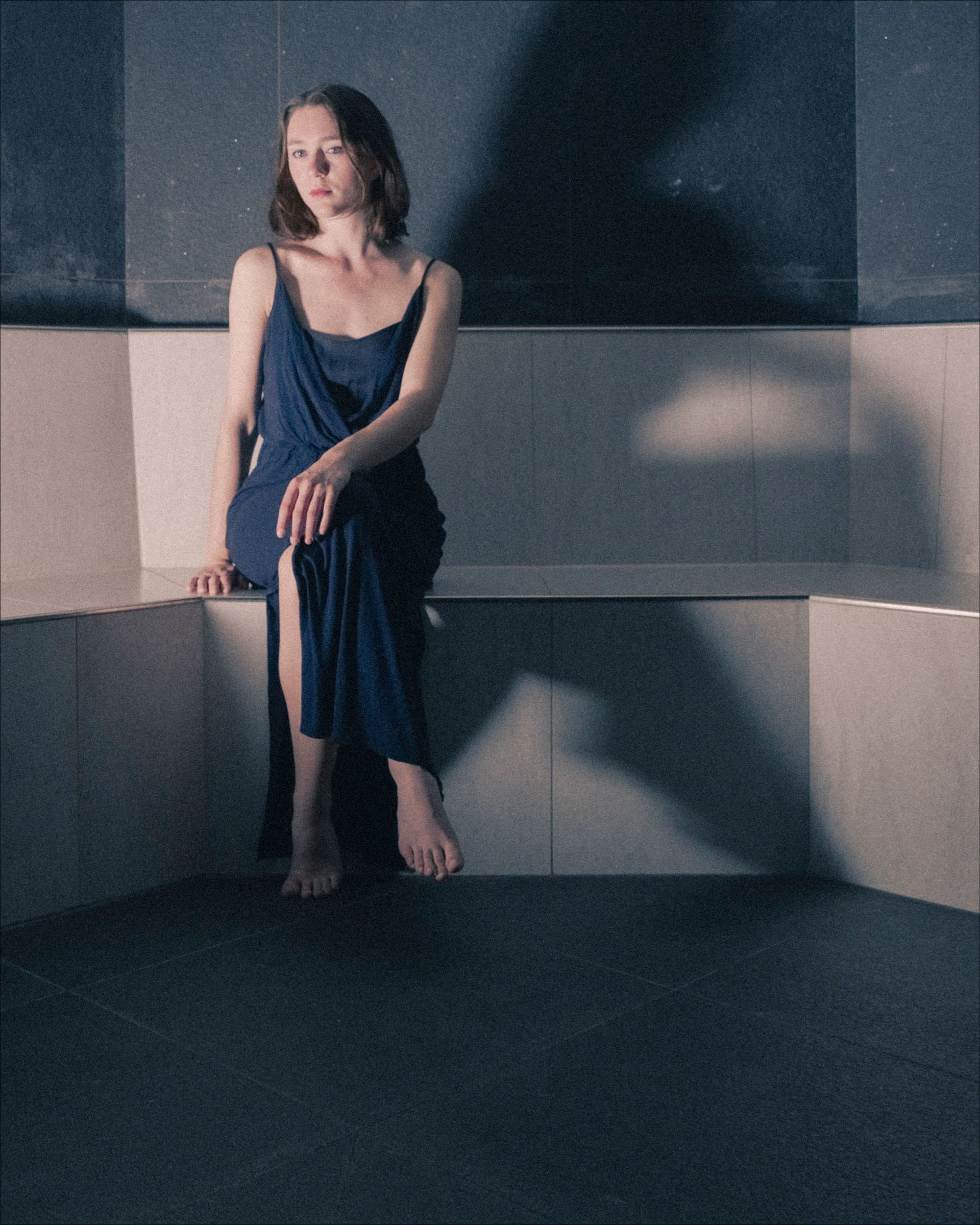 Woman in blue dress sits on a white ledge in a dimly lit, dark space, with a large shadow behind her.
