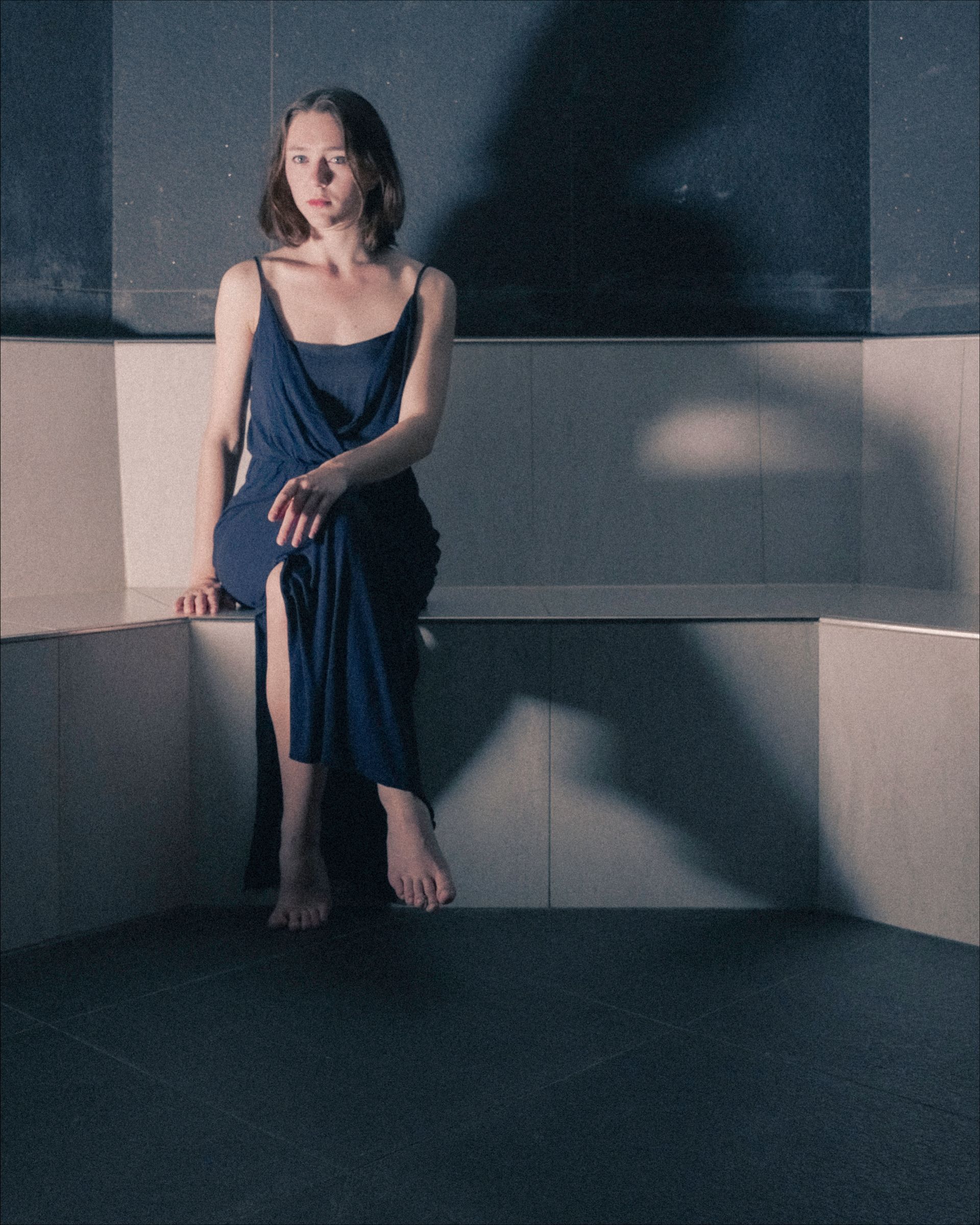 Woman in blue dress sits on ledge, looking away, with shadow on wall.