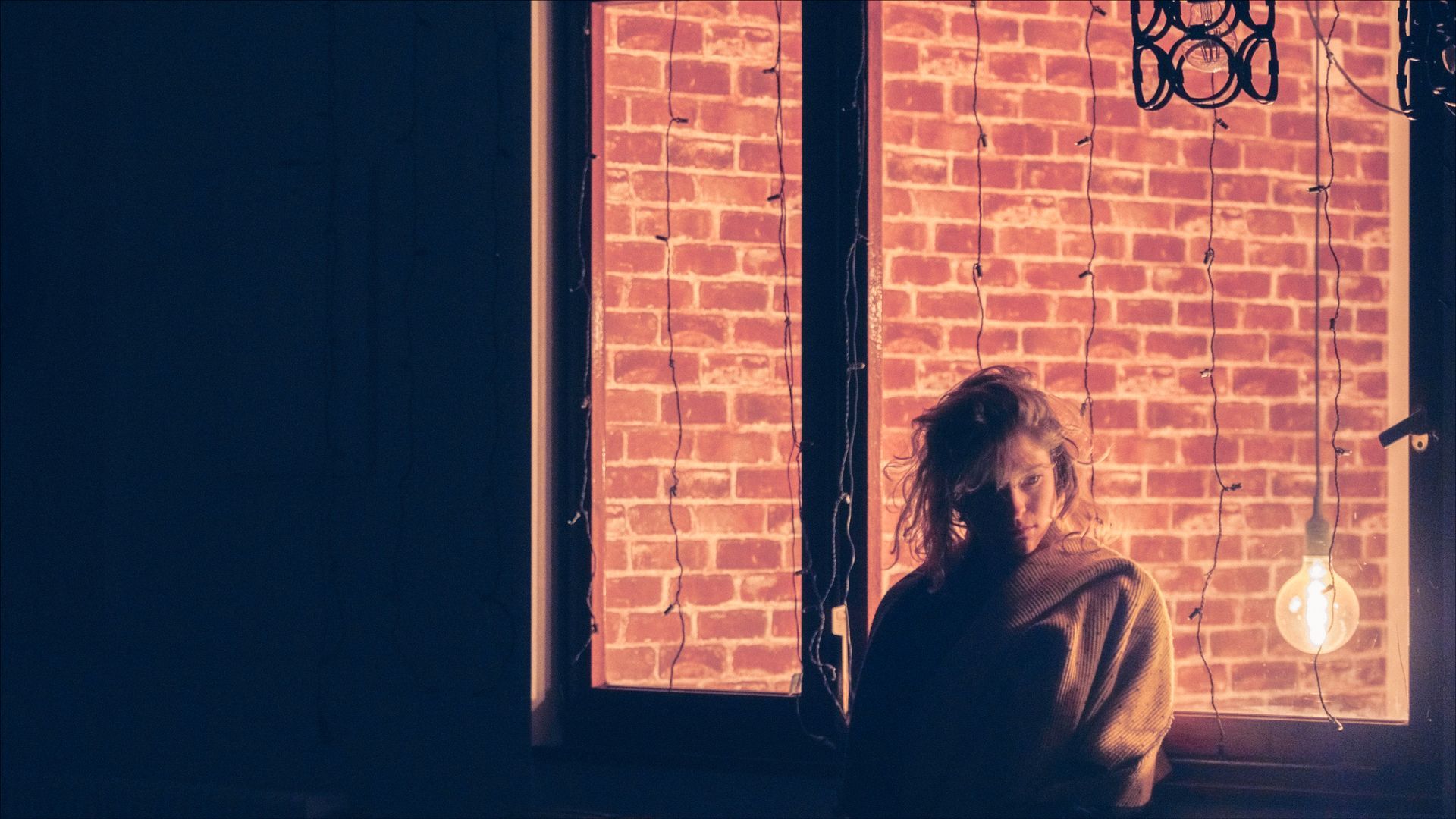 Woman standing by window; brick wall background, lit bulb hanging. Dark interior with orange hues.