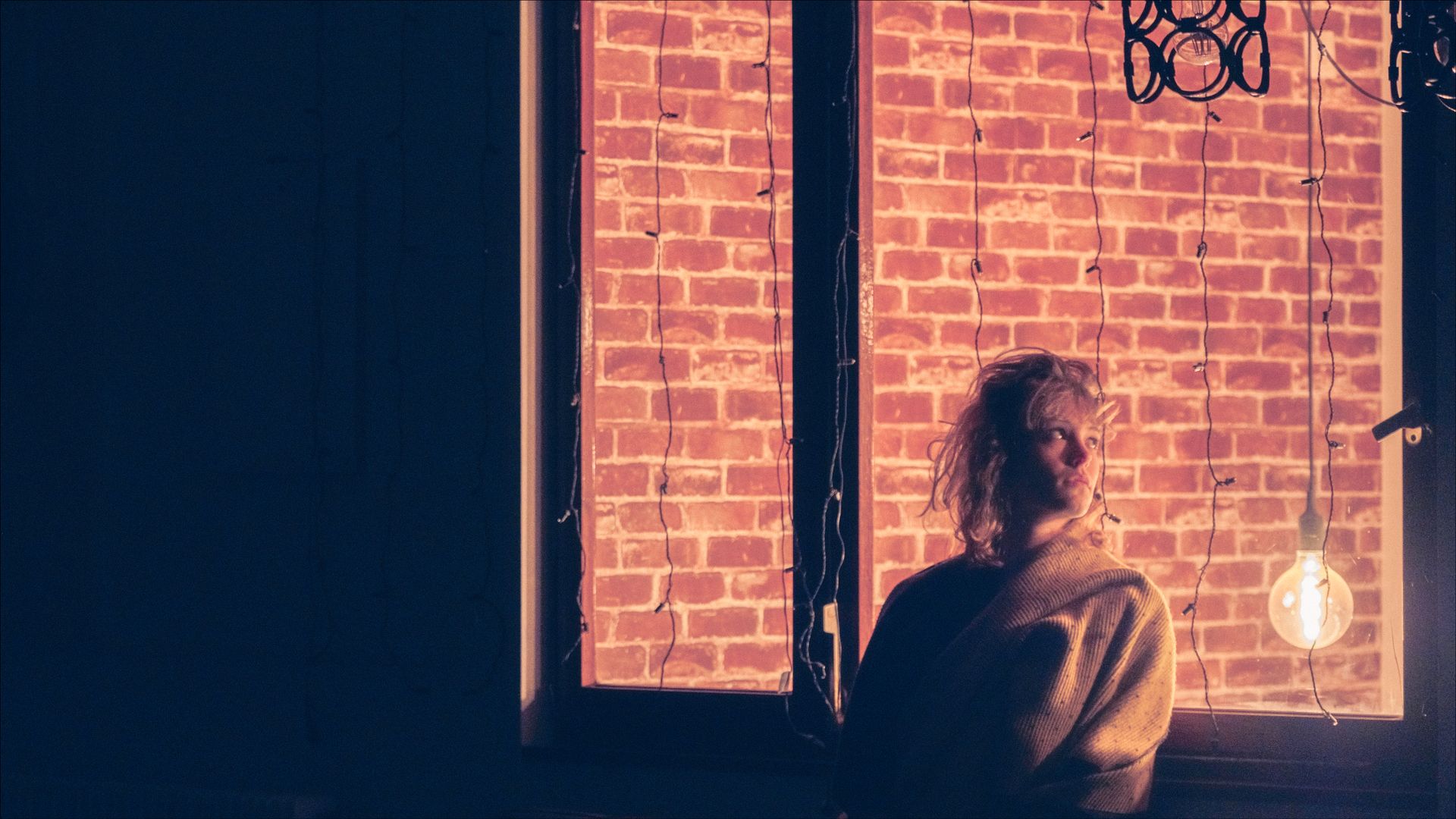 Person in front of a brick window, lit by a hanging bulb. Dark interior, warm tones.