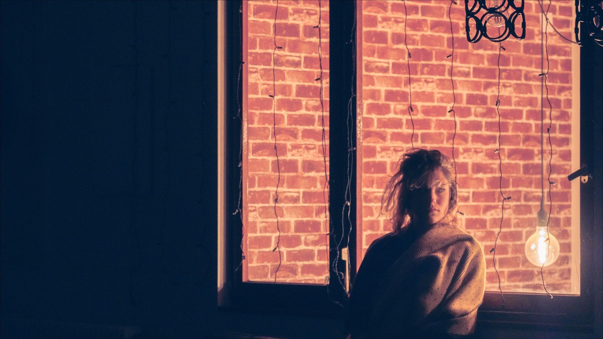 Woman standing by a window with a brick wall visible behind her, lit by a warm light.