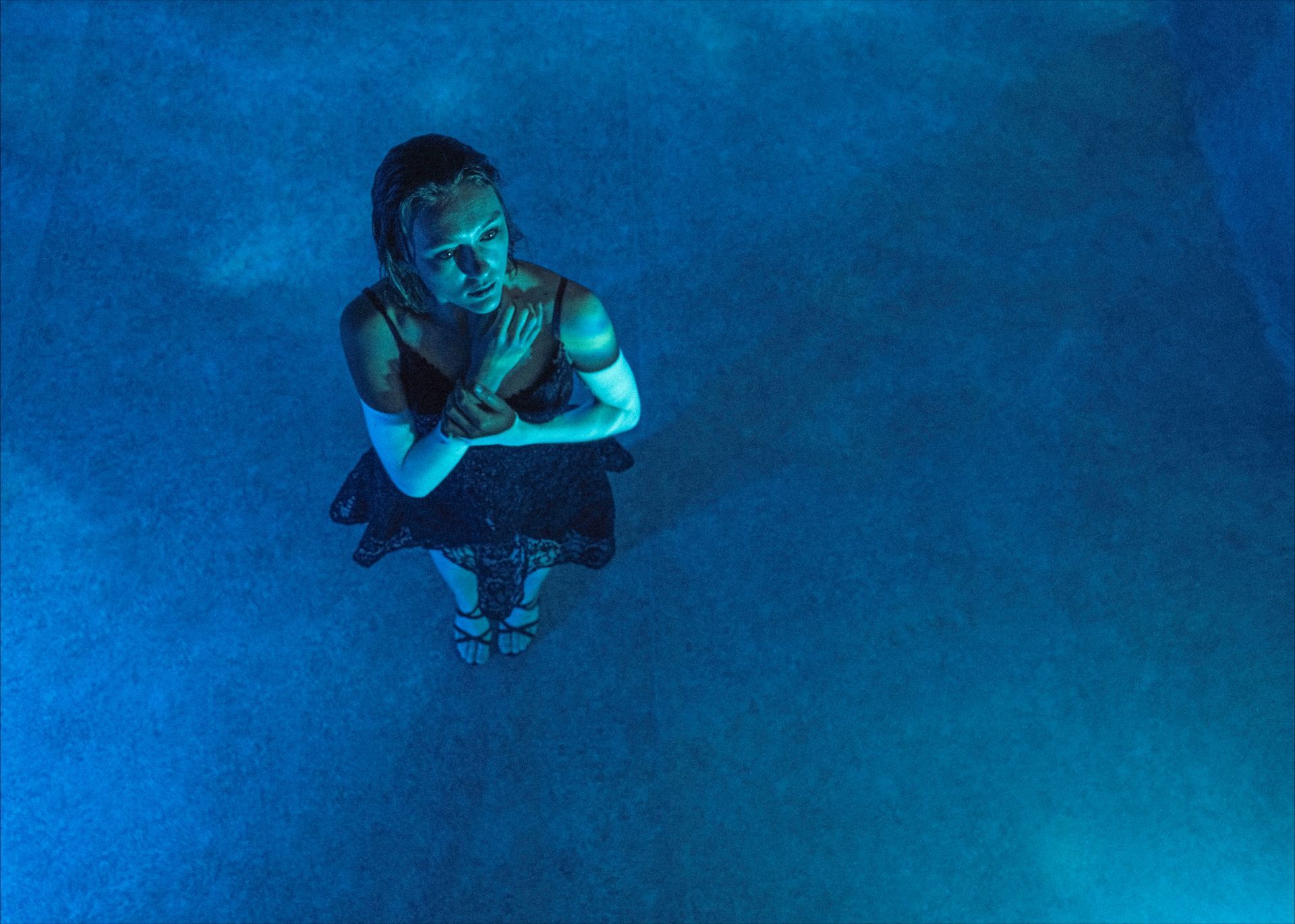 Woman in a black dress stands in blue-lit mist, arms crossed, looking upward.