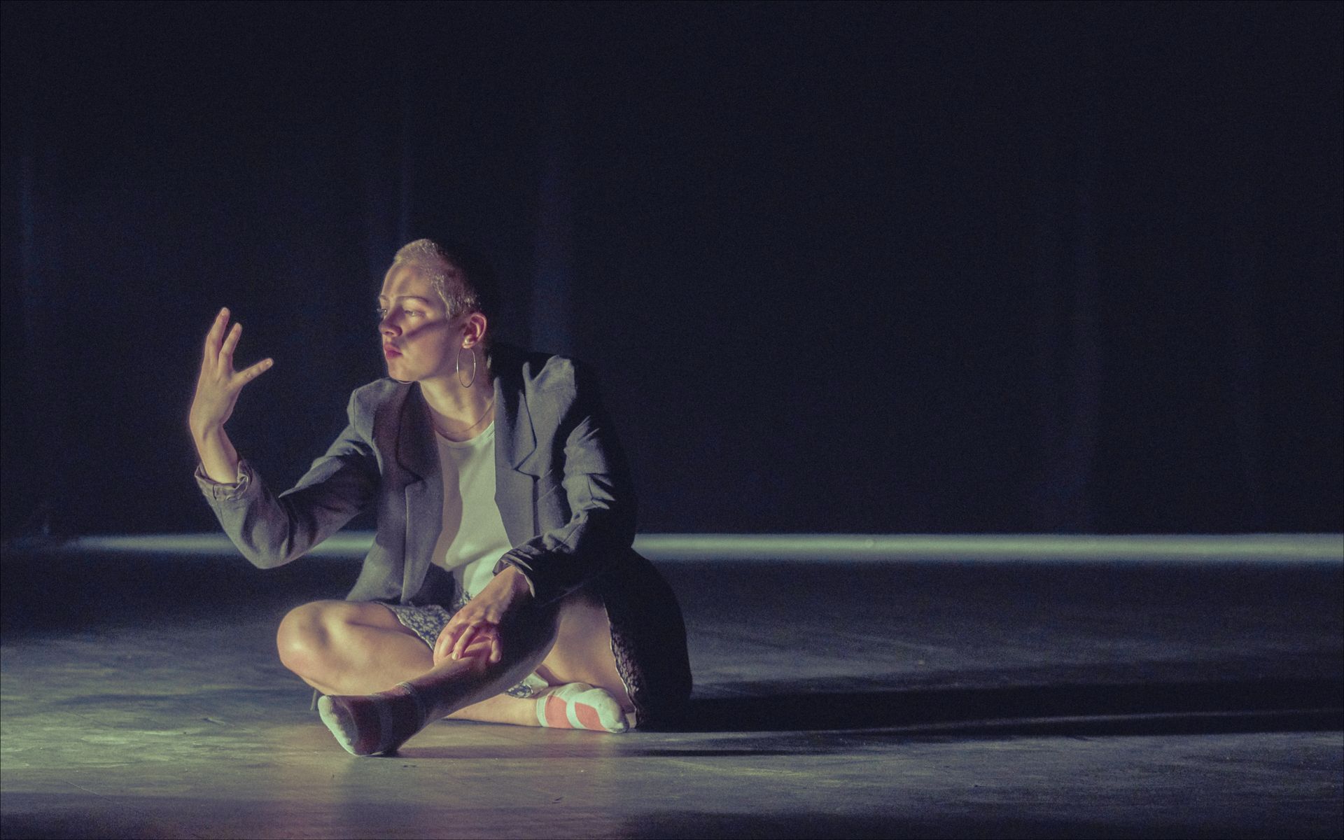 Person in blazer sits cross-legged, hand raised, lit from above, in a dark space.