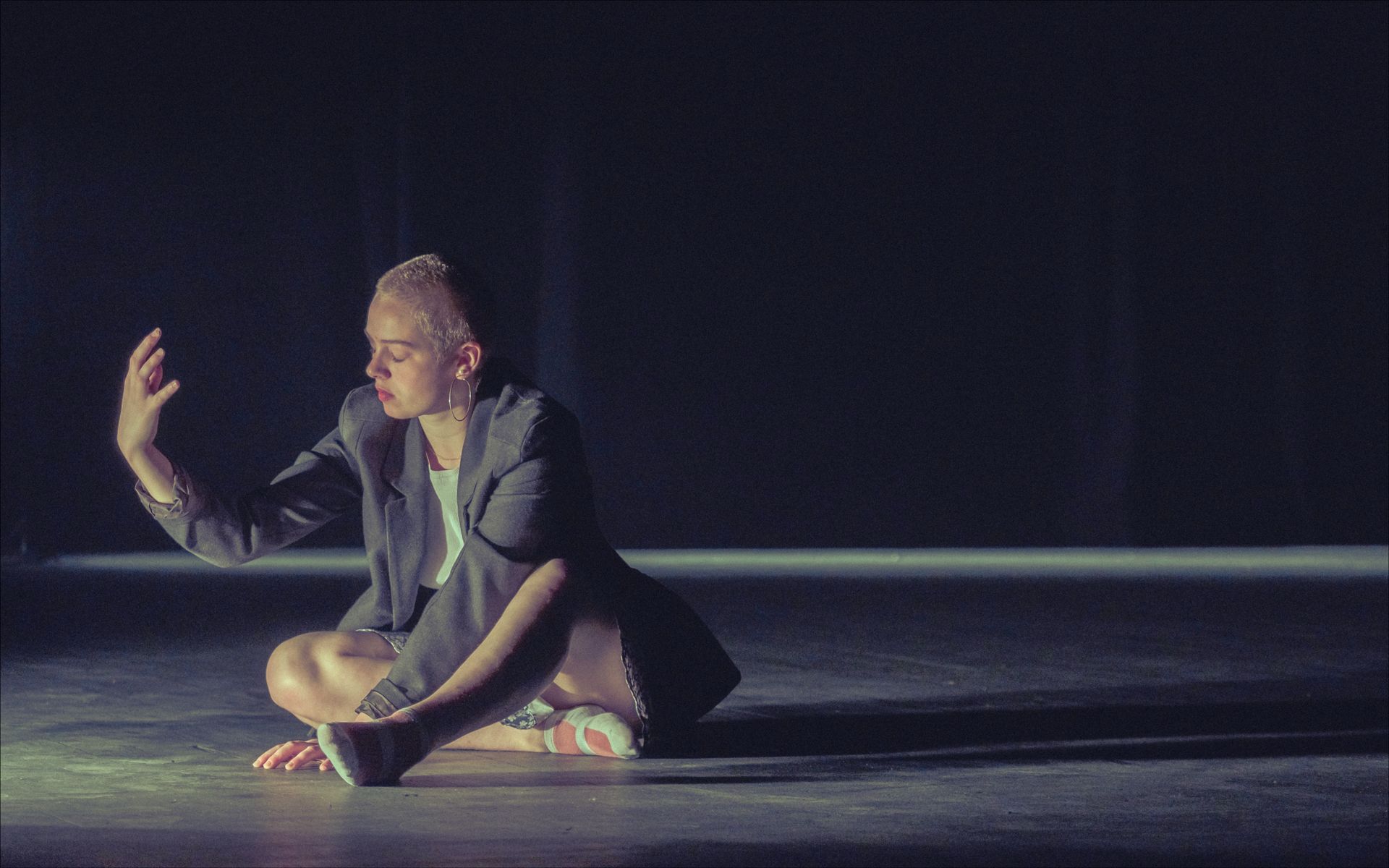 Dancer in a blazer, seated cross-legged on a dark stage, arm raised towards light.