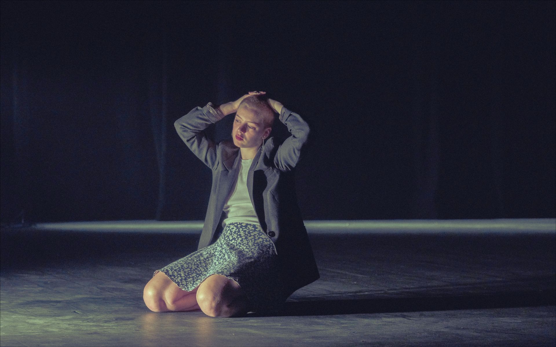 Person on knees, hands on head, in a dark space, lit by a spotlight.