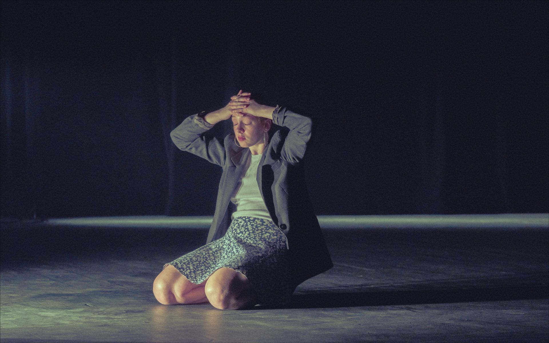 Person kneels on stage, hands on head, eyes closed. Dark room, spotlight.