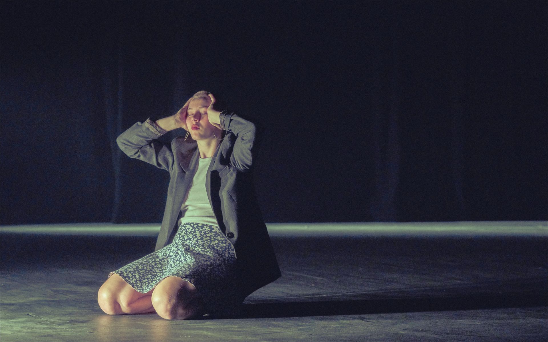 Person on knees, hands on head, on a dark stage. Wearing a jacket, t-shirt, and patterned shorts.